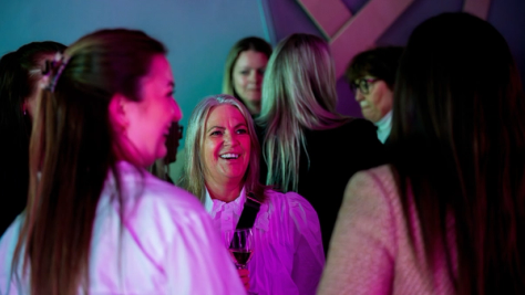 En smilende kvinne med vinglass omgitt av andre kvinner i rosa belysning på et nettverksarrangement — av Nonspace