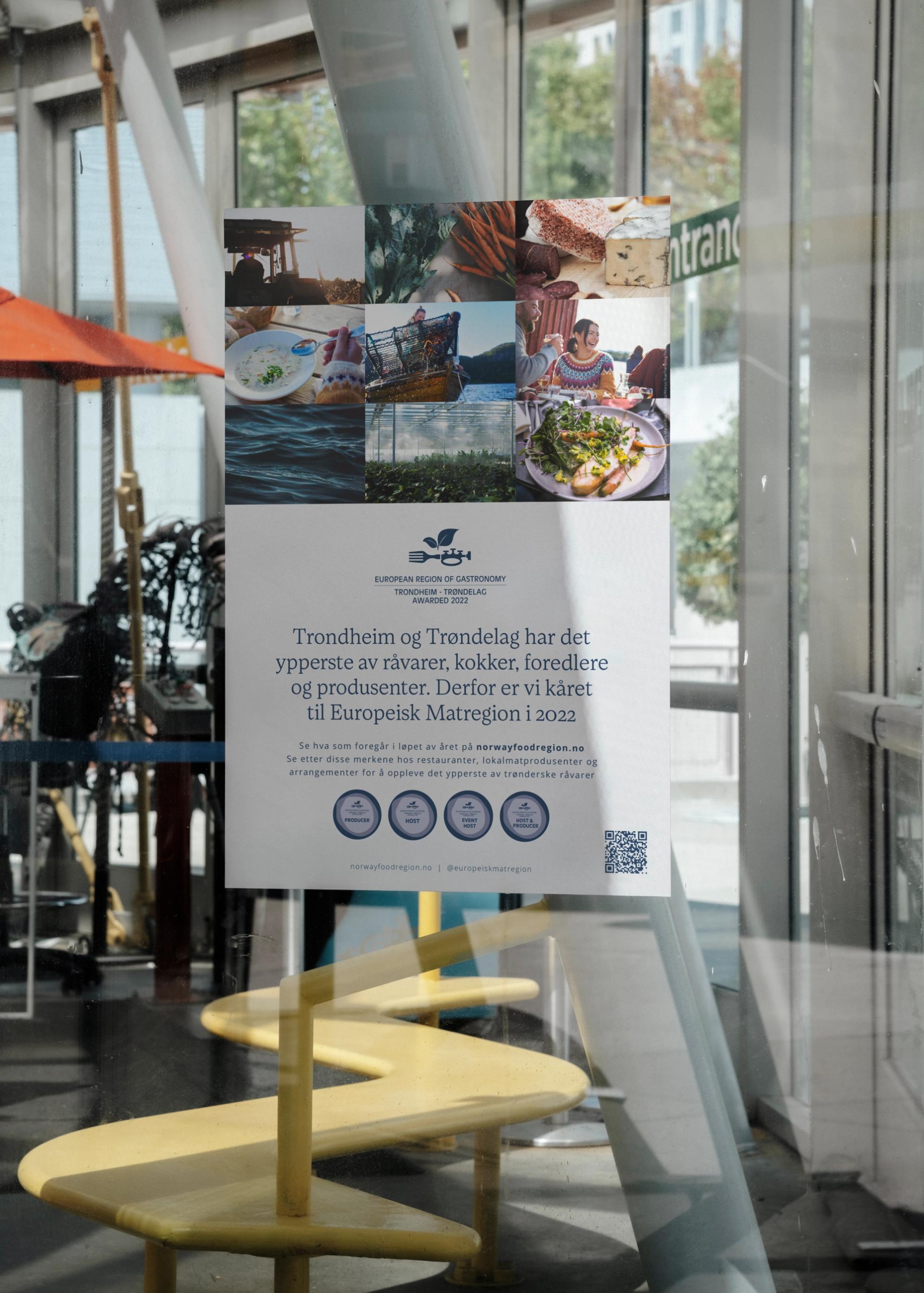 Plakat om Trondheim-Trøndelag som Europeisk Matregion 2022 hengt opp i et vindu med matbilder og QR-kode — av Nonspace