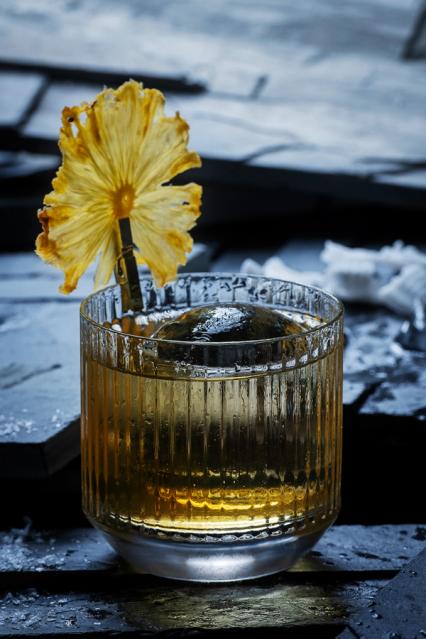 A whiskey cocktail in a ribbed glass garnished with a dried pineapple slice on a dark slate surface — by Nonspace