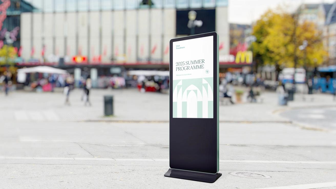 En digital reklameskjerm på et bytorg viser Visit Trondheims 2025 Summer Programme med katedral-grafikk — av Nonspace