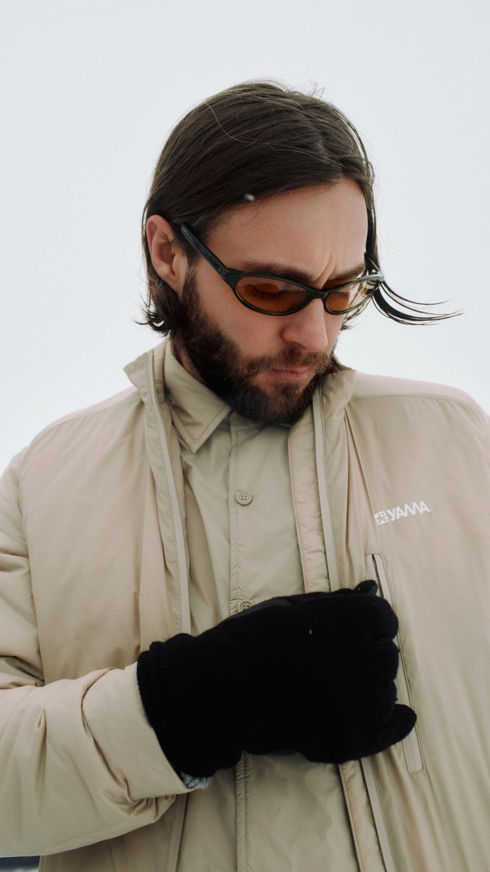 En skjeggete mann med solbriller og beige jakke ser ned mens han holder hendene foran seg med svarte hansker — av Nonspace