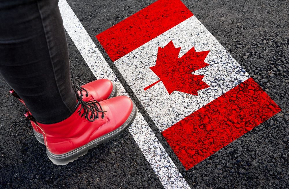 A person wearing red boots standing at a painted border line featuring the Canadian flag on the asphalt, symbolizing entry into Canada with a criminal record.