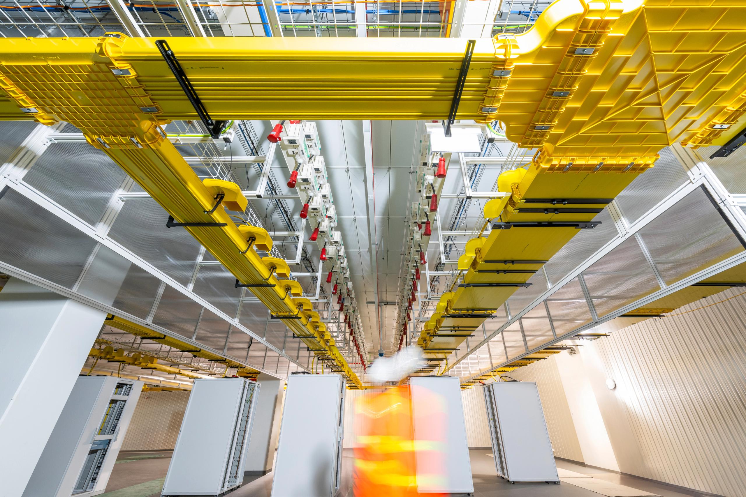 Yellow overhead cable trays running the length of a data hall ceiling, with a worker in motion blur below