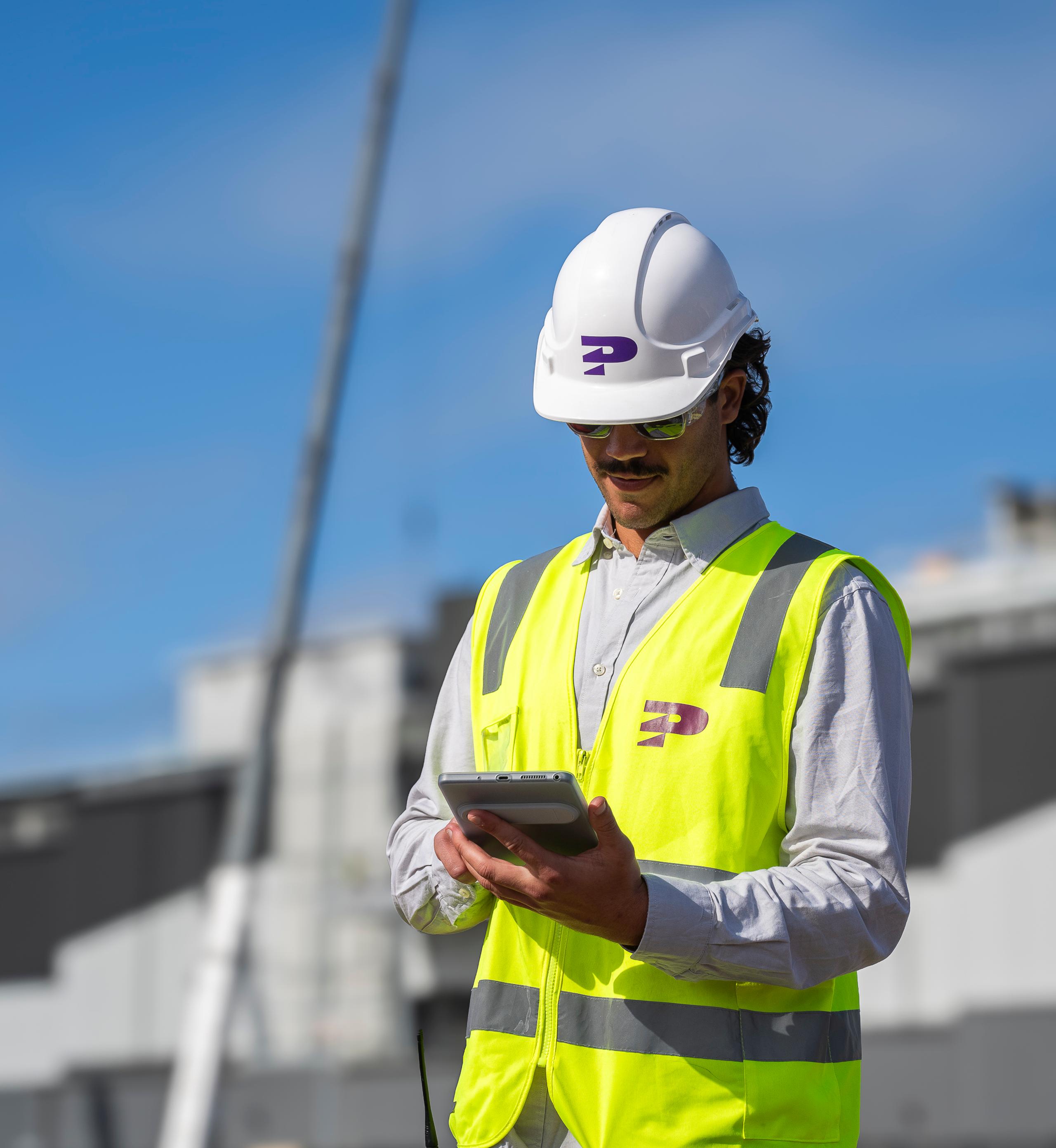 A Pinnacl worker in hi-vis and hard hat using a tablet on a construction site, with a crane behind
