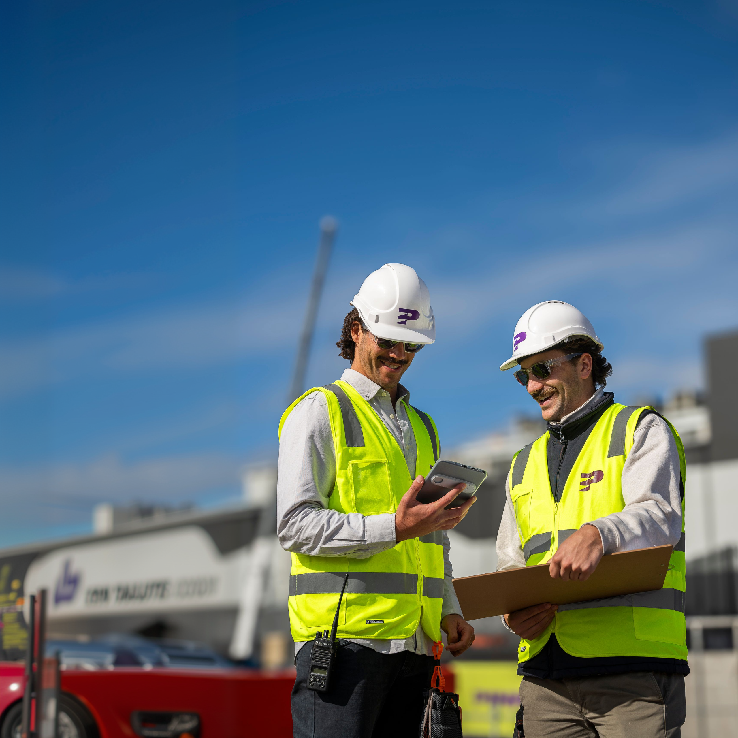 Two Pinnacl workers in hi-vis vests and hard hats smiling at a tablet on a sunny construction site