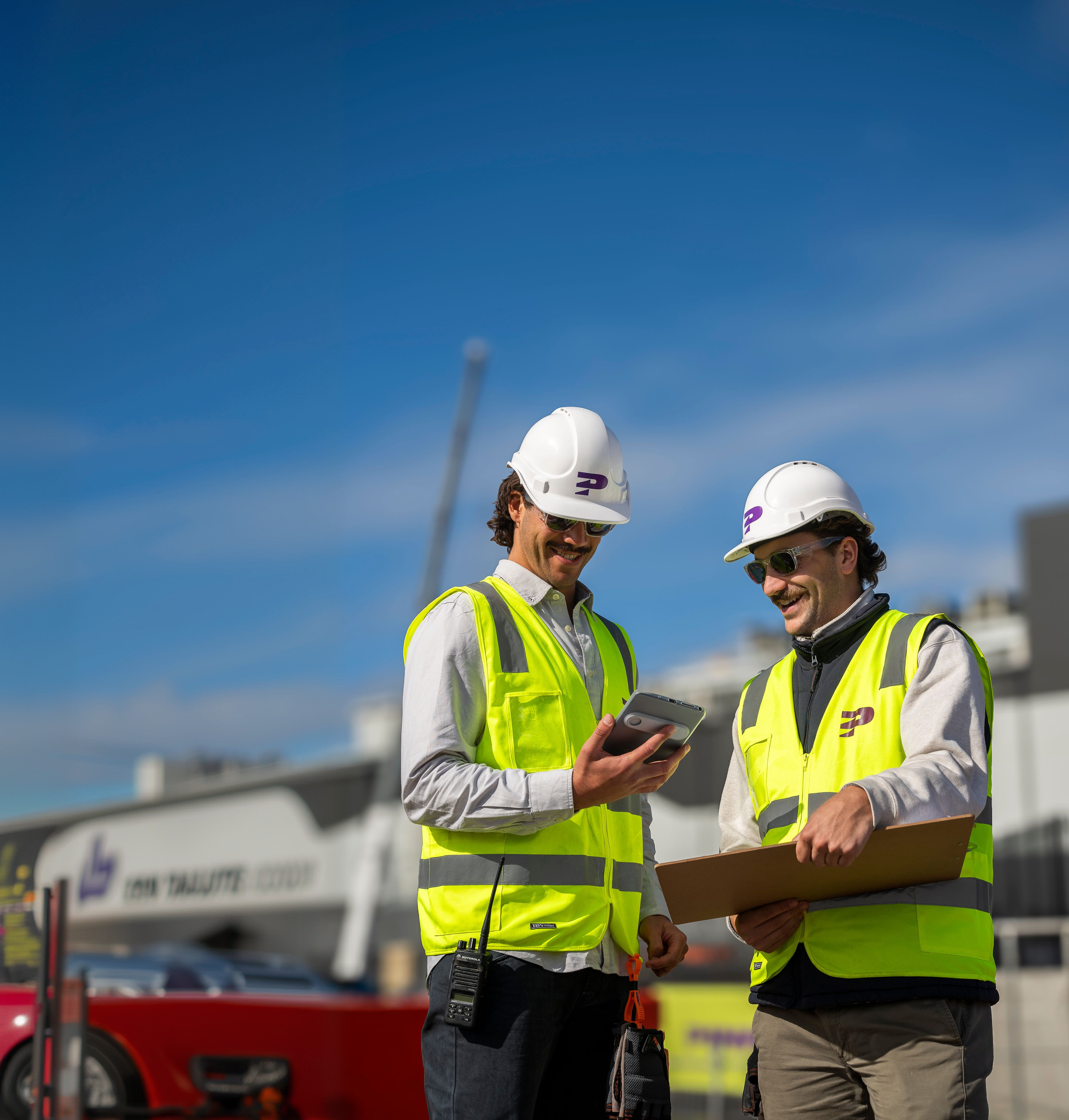 Two Pinnacl workers in hi-vis vests and hard hats smiling at a tablet on a sunny construction site