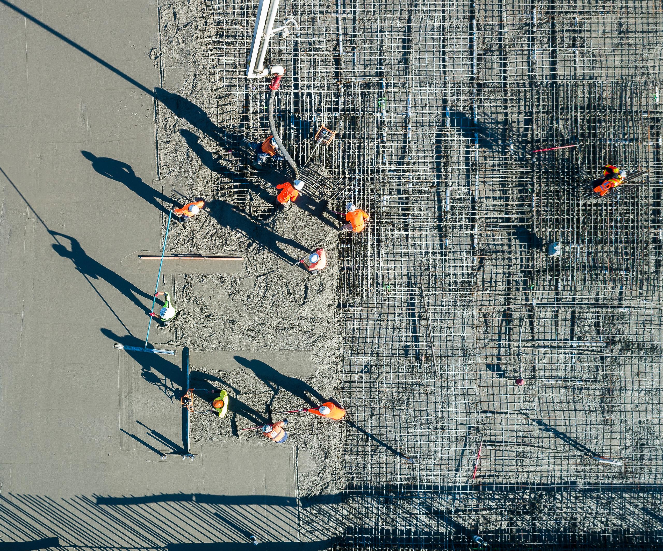 Aerial top-down view of workers placing concrete on a reinforced slab, casting long shadows