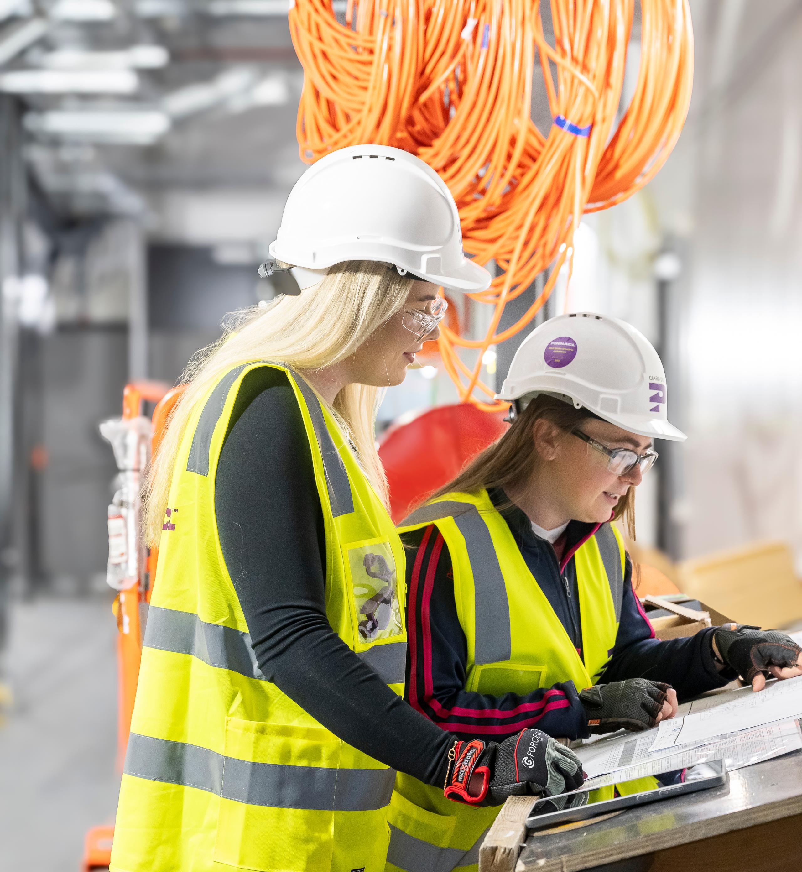 Two female Pinnacl workers in hi-vis and hard hats reviewing plans at a workbench beneath coiled orange cabling