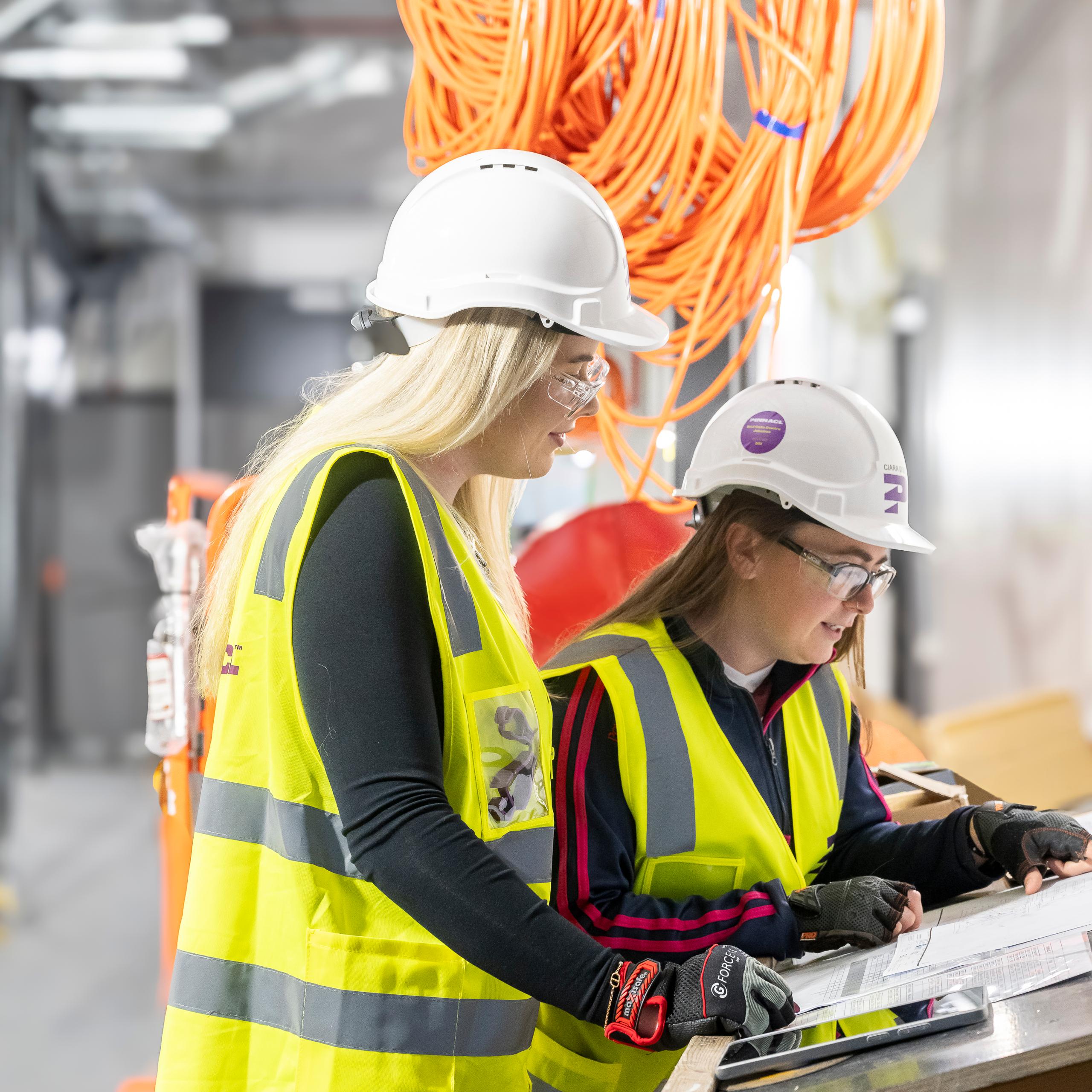 Two female Pinnacl workers in hi-vis and hard hats reviewing plans at a workbench beneath coiled orange cabling