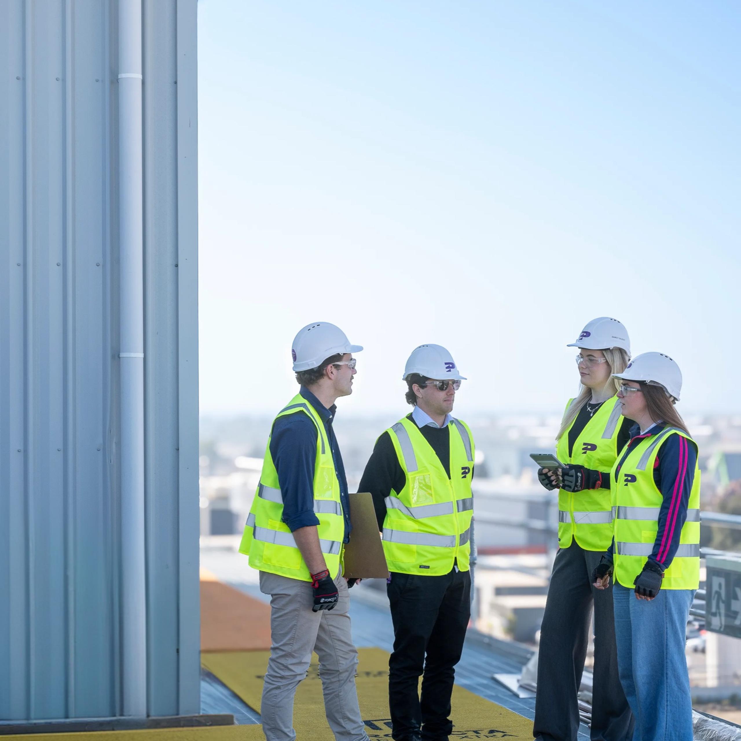 Four Pinnacl workers in hi-vis and hard hats talking on a rooftop deck with the city skyline behind