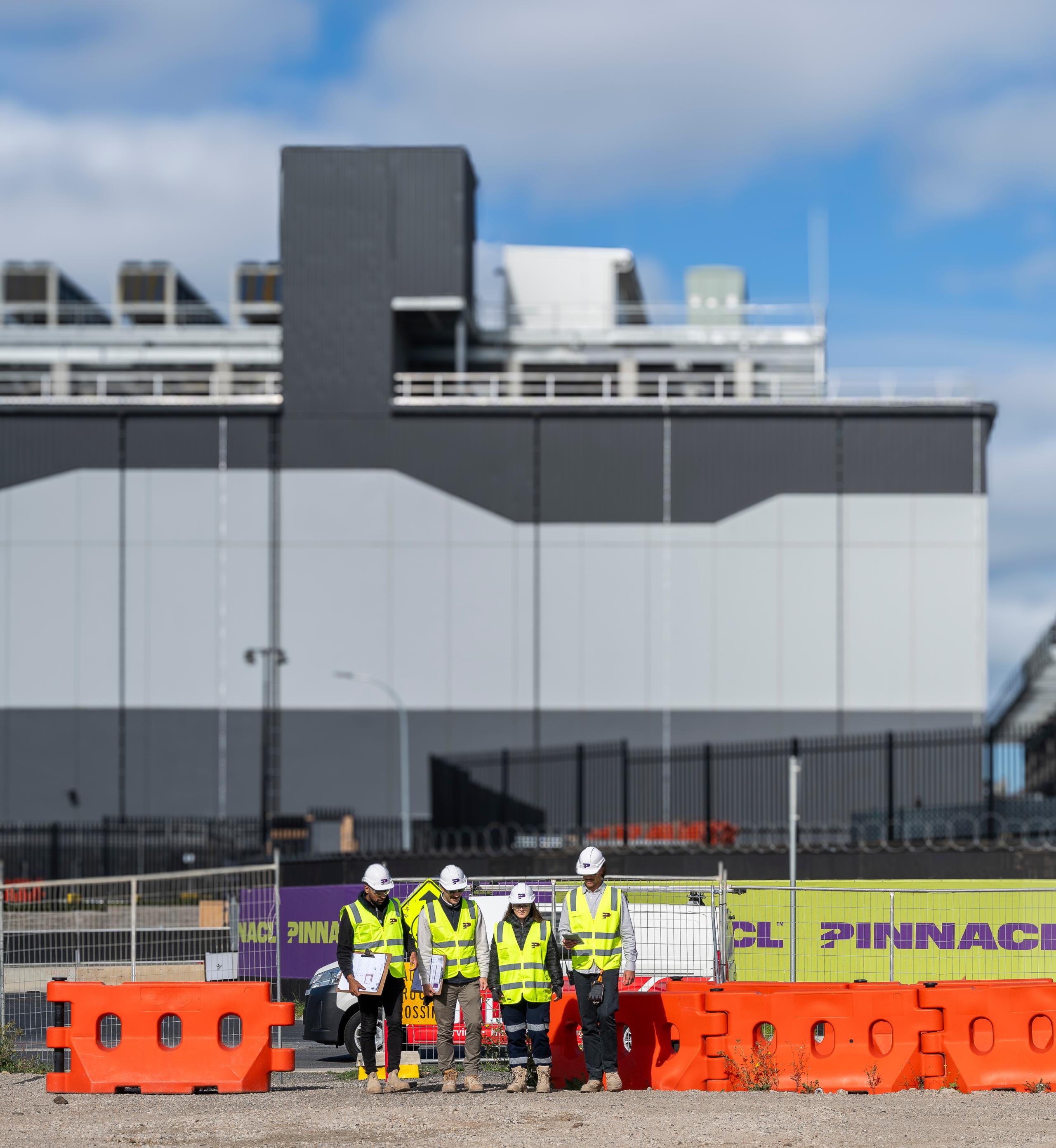 Exterior of a large grey data centre building with Pinnacl workers behind orange site barriers in the foreground