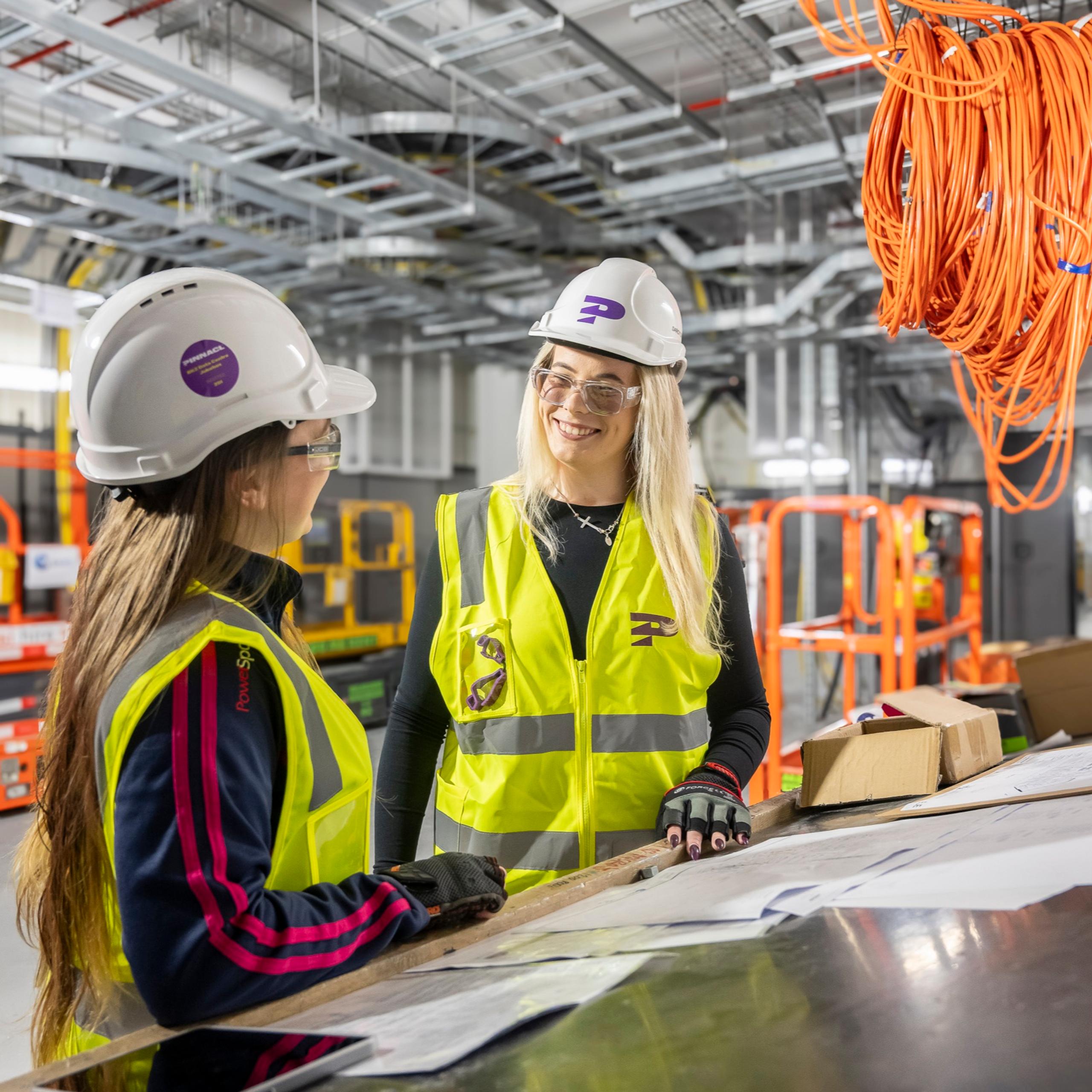 Two female Pinnacl workers in hi-vis and hard hats talking across a bench inside a building under fit-out