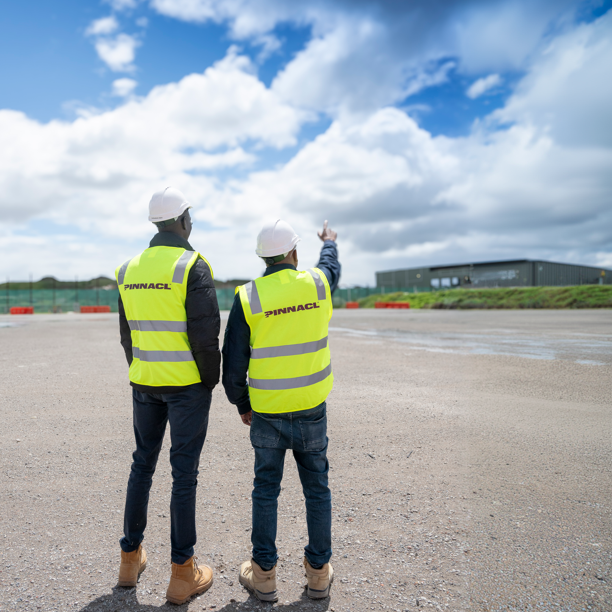 Two Pinnacl workers in hi-vis vests and hard hats looking across an open construction site, one pointing into the distance