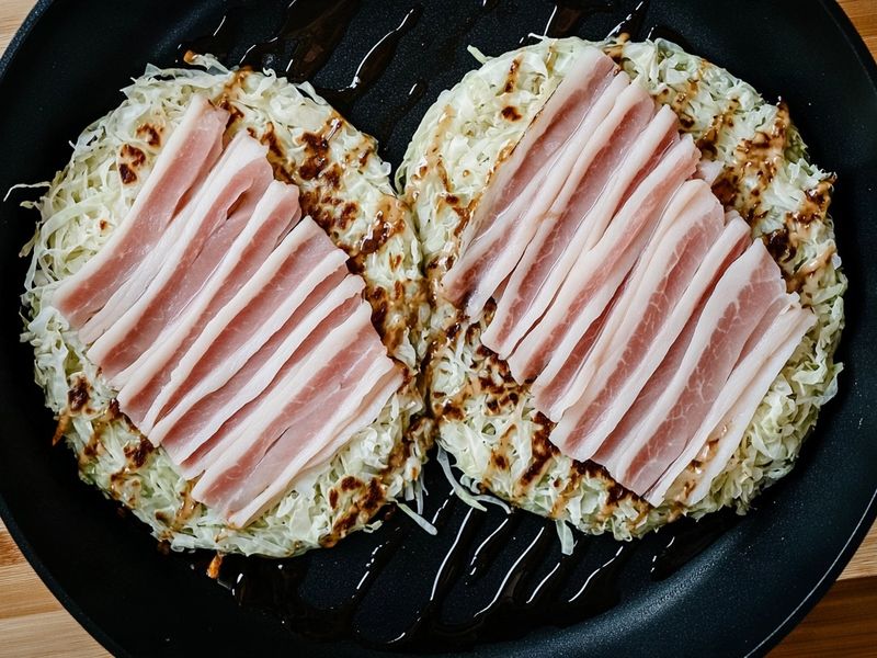 Two cabbage pancakes in a skillet topped with raw pork belly slices before flipping