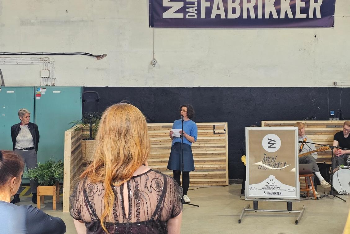 Bilde av en kvinne som prater i mikrofon foran en liten gruppe mennesker. Det står Nydalen Fabrikker på en plakat bak henne.