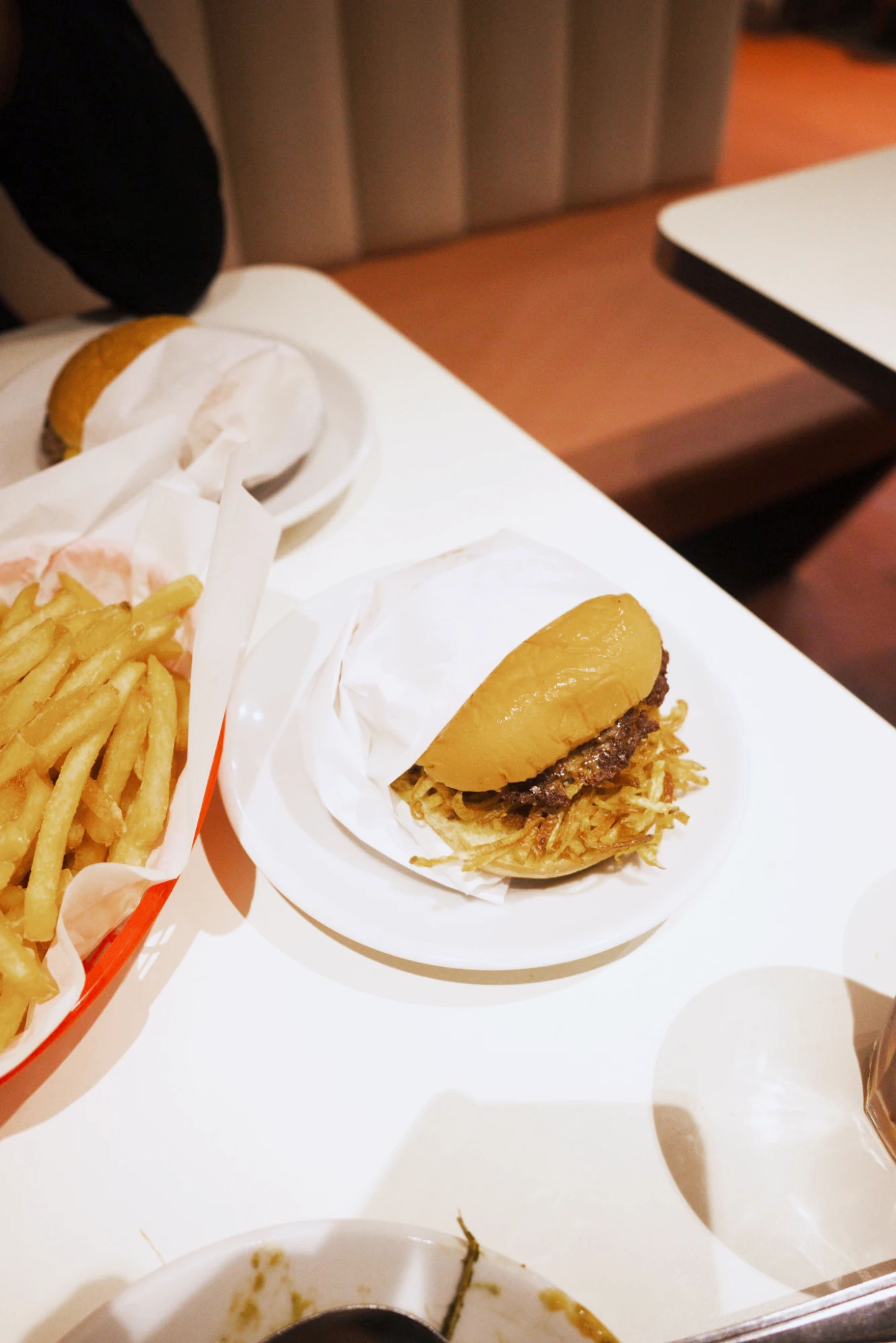 Lima Kreuzburger mit Kartoffelstäbchen unter dem Patty auf einem weißen Teller, daneben ein roter Korb mit Pommes