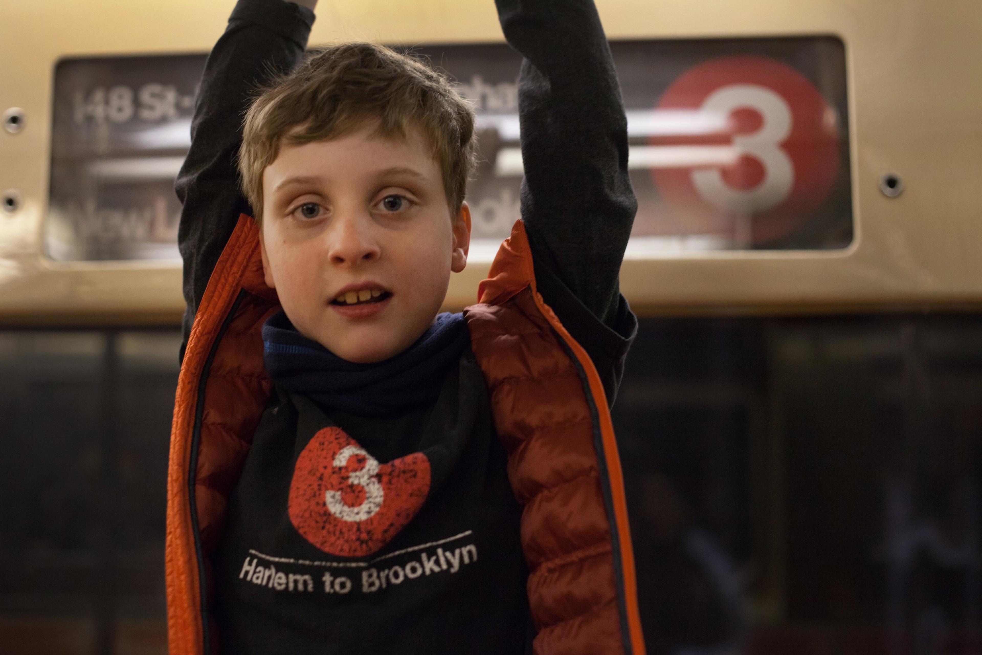 Colin raises his arms under a 3 train destination sign, wearing a T-shirt with a 3 on it.