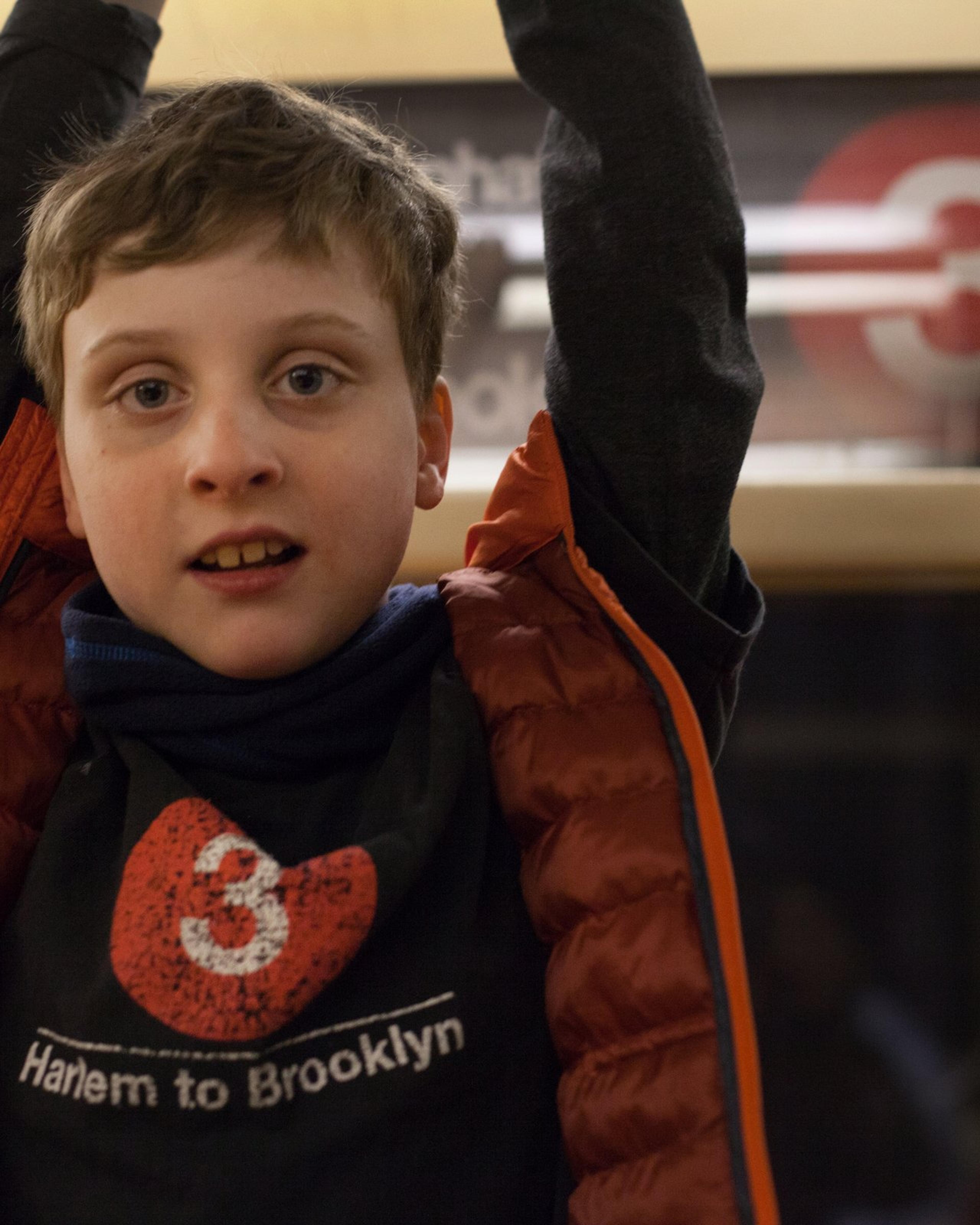 Colin raises his arms under a 3 train destination sign, wearing a T-shirt with a 3 on it.