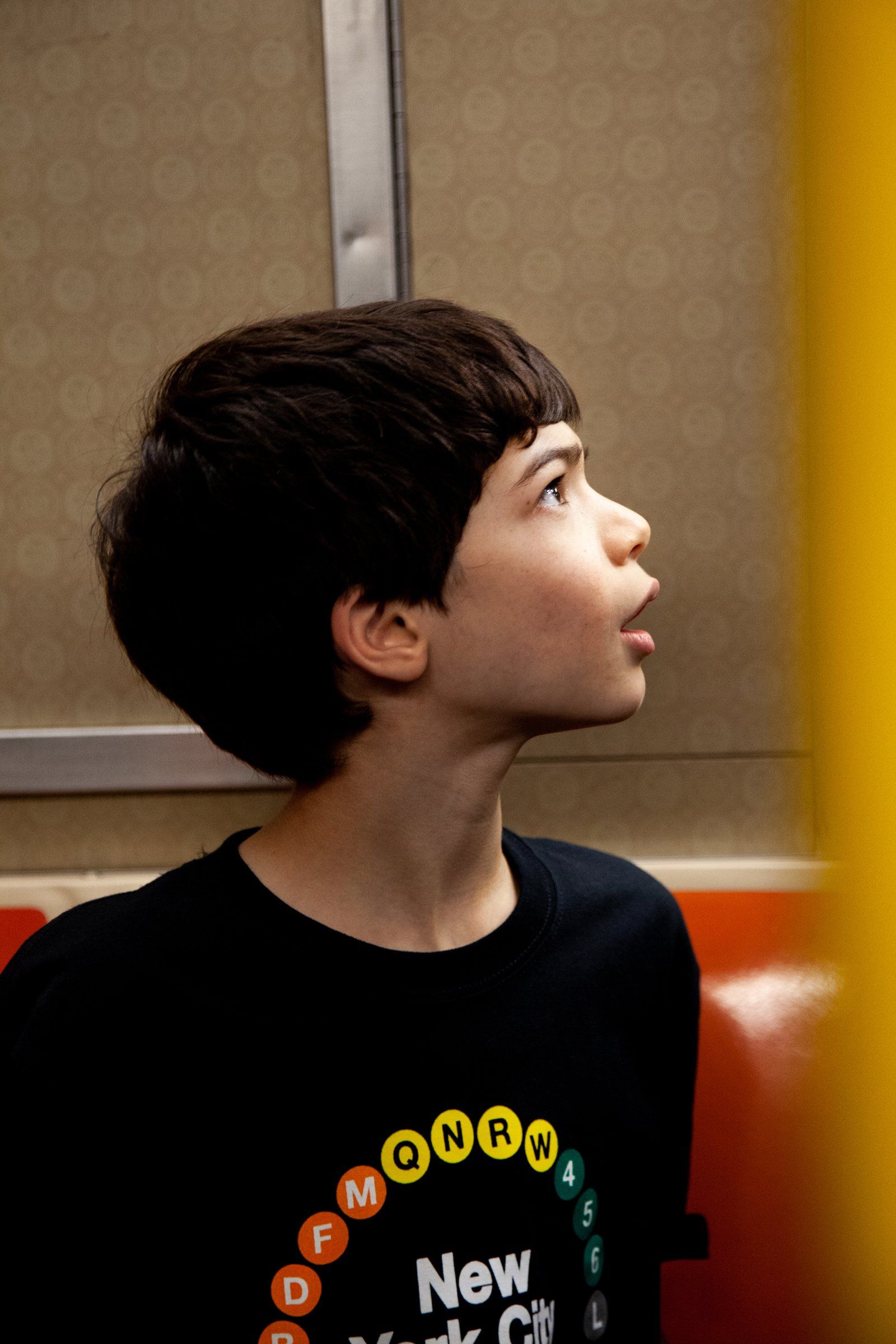 Jake, in a T-shirt printed with subway line bullets, gazes up inside a subway car.