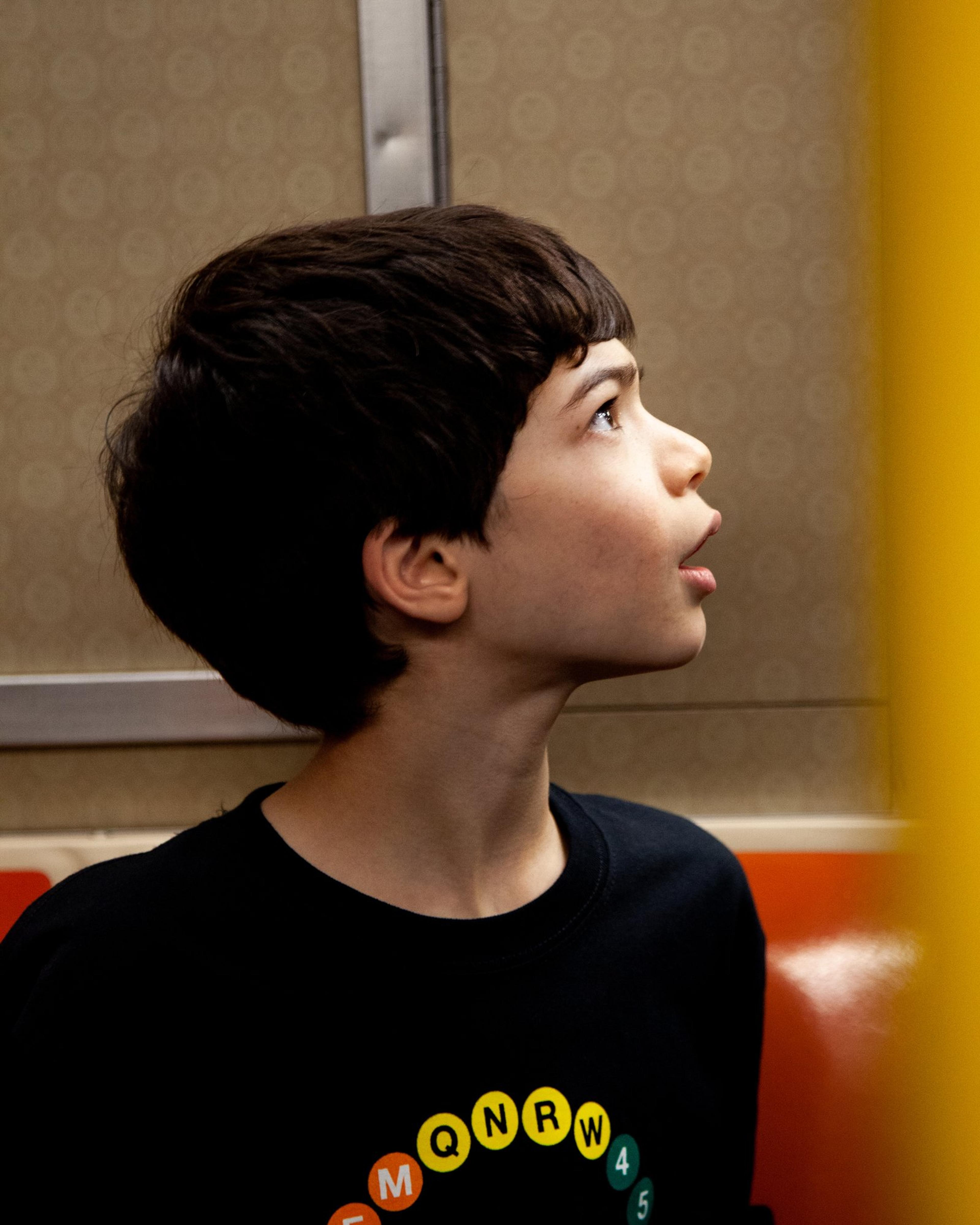 Jake, in a T-shirt printed with subway line bullets, gazes up inside a subway car.
