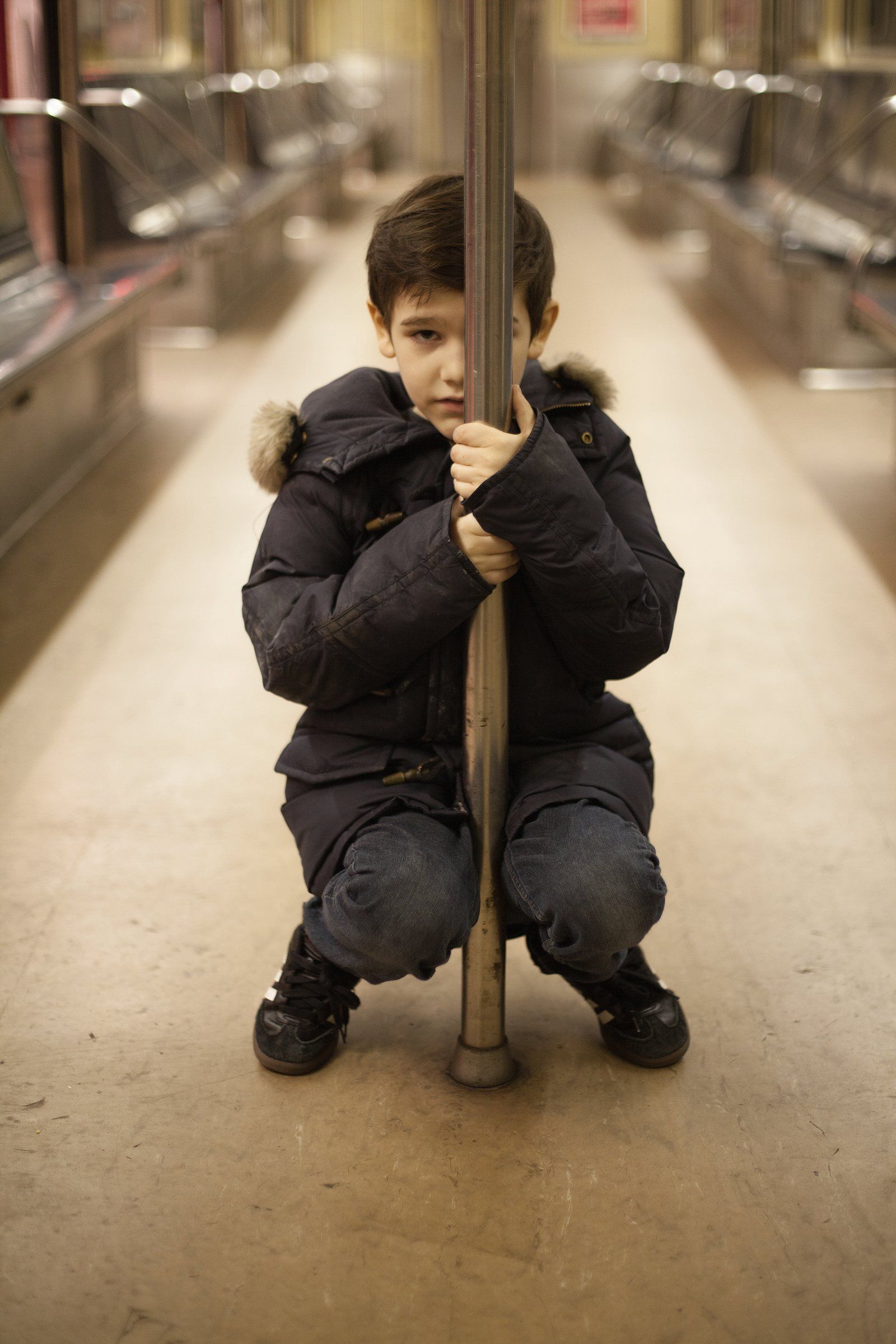 Orson, in a dark winter coat, crouches on the floor of an empty subway car, hugging a grab pole.