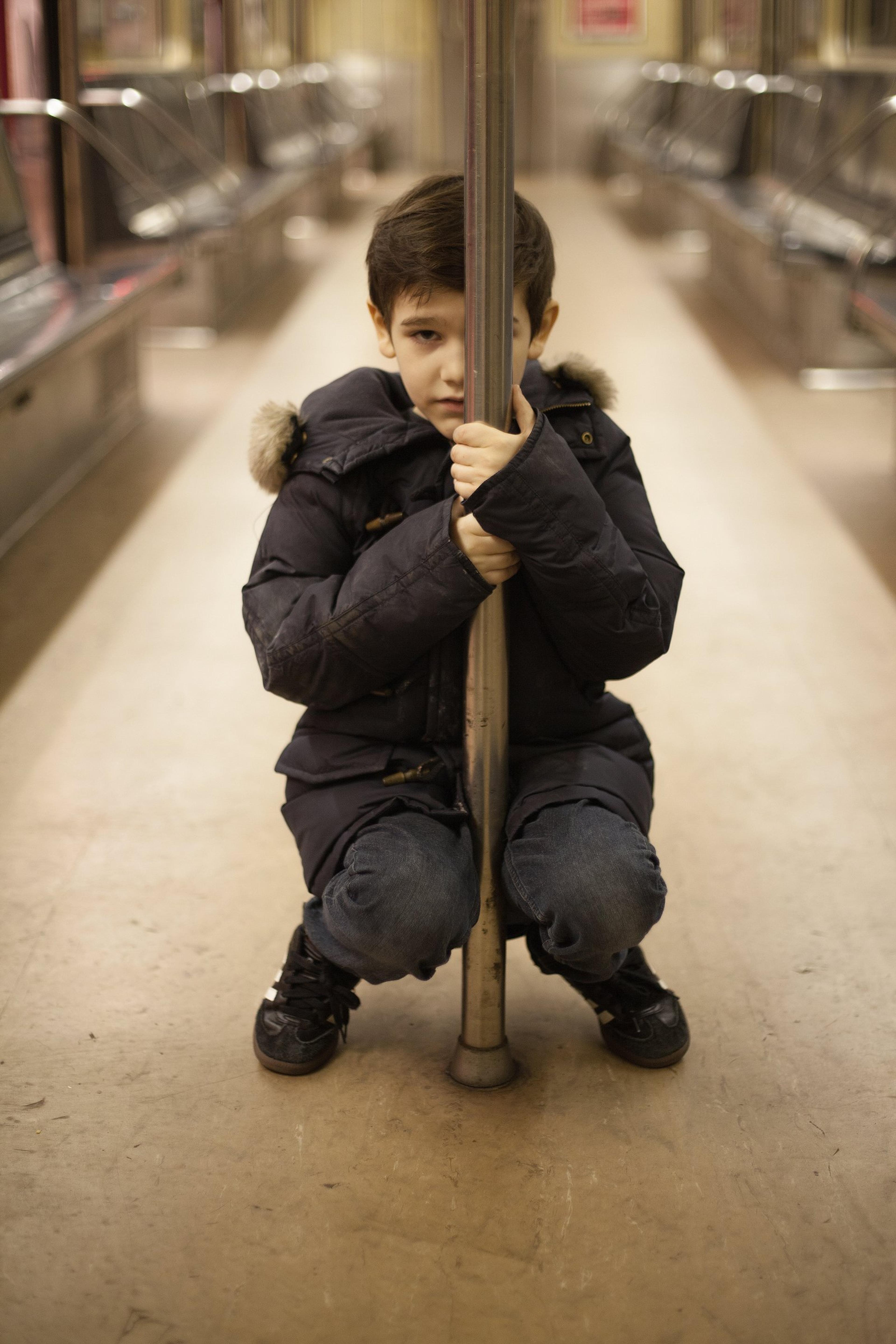 Orson, in a dark winter coat, crouches on the floor of an empty subway car, hugging a grab pole.