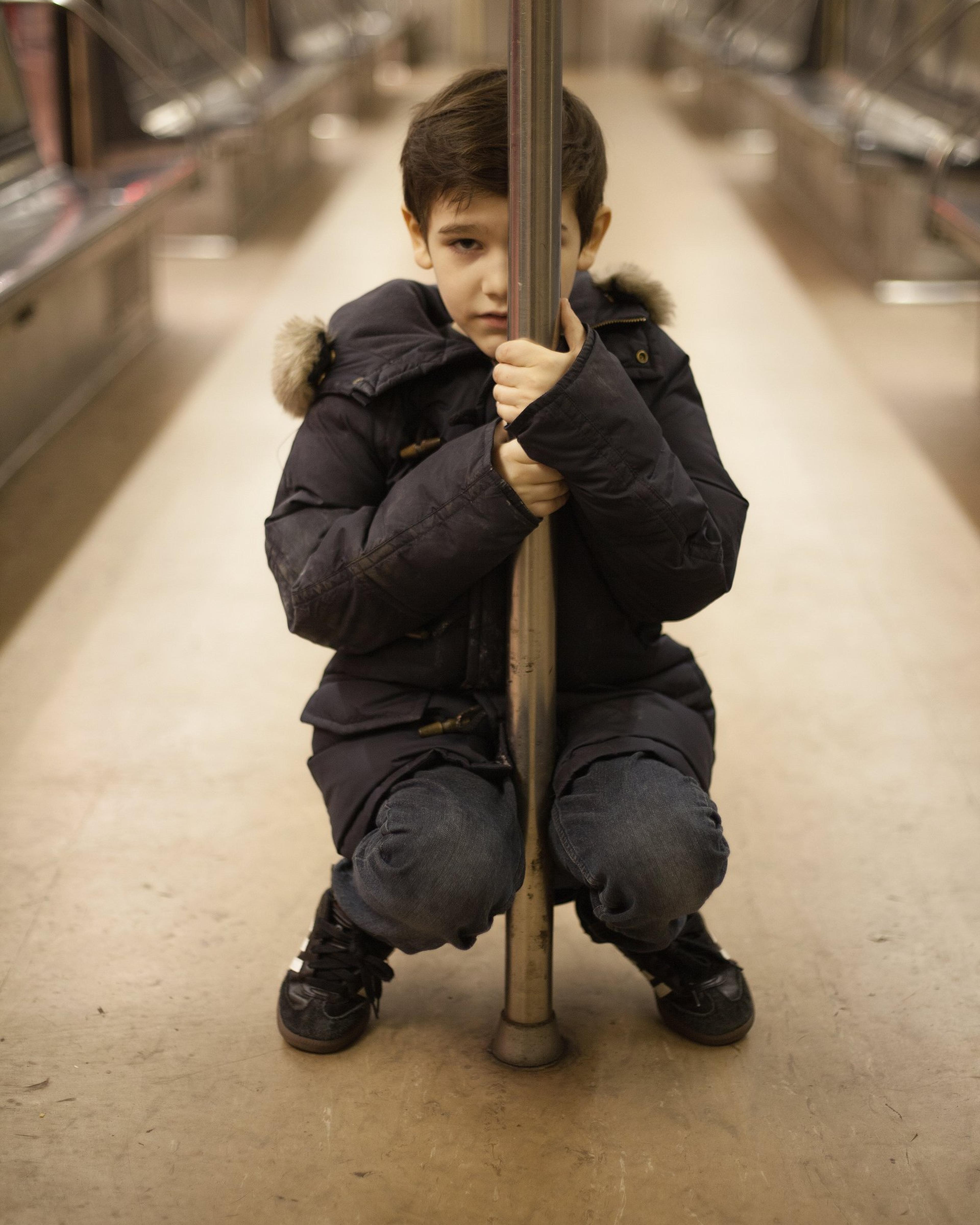 Orson, in a dark winter coat, crouches on the floor of an empty subway car, hugging a grab pole.