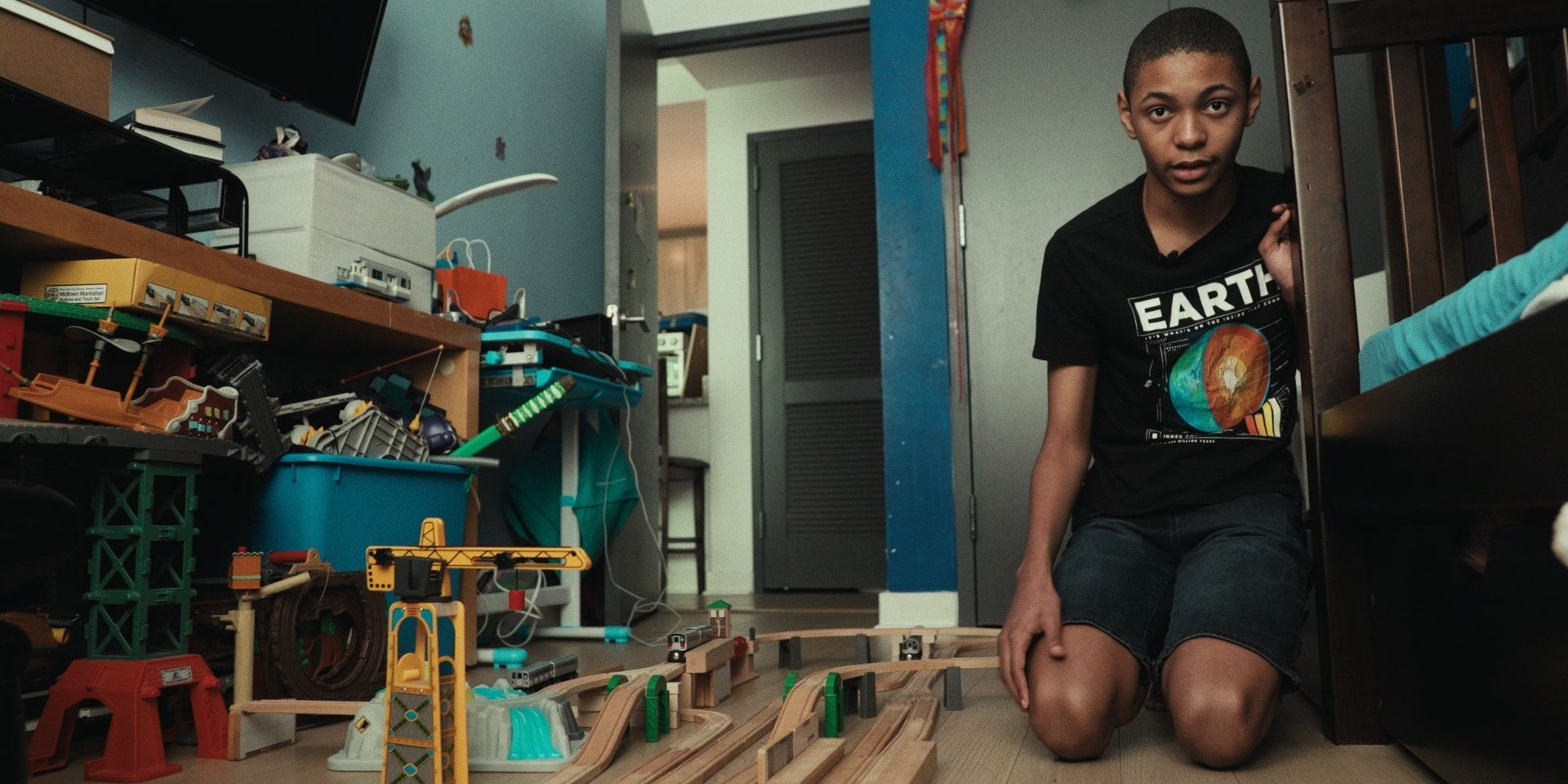 A boy kneels among the wooden train tracks spread across his bedroom floor.