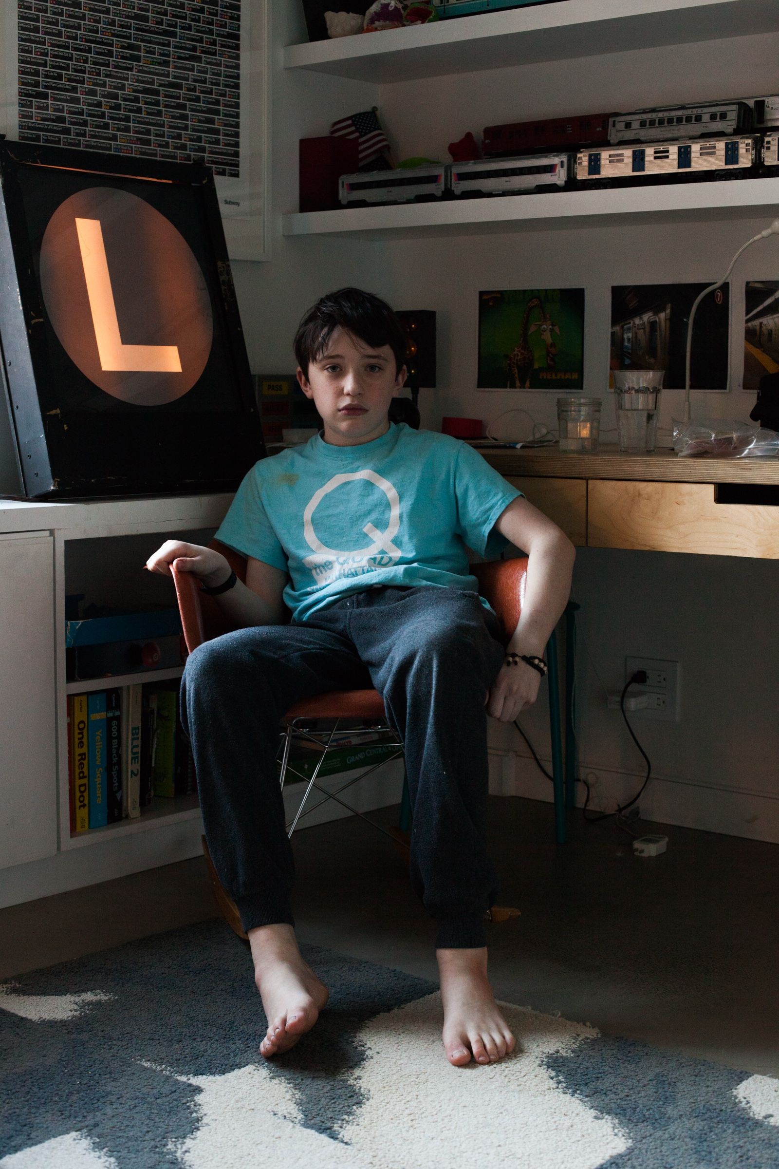 Booker, barefoot in a blue T-shirt, sits in a chair in his room beside a lit-up L train sign, model trains lining the shelves behind him.