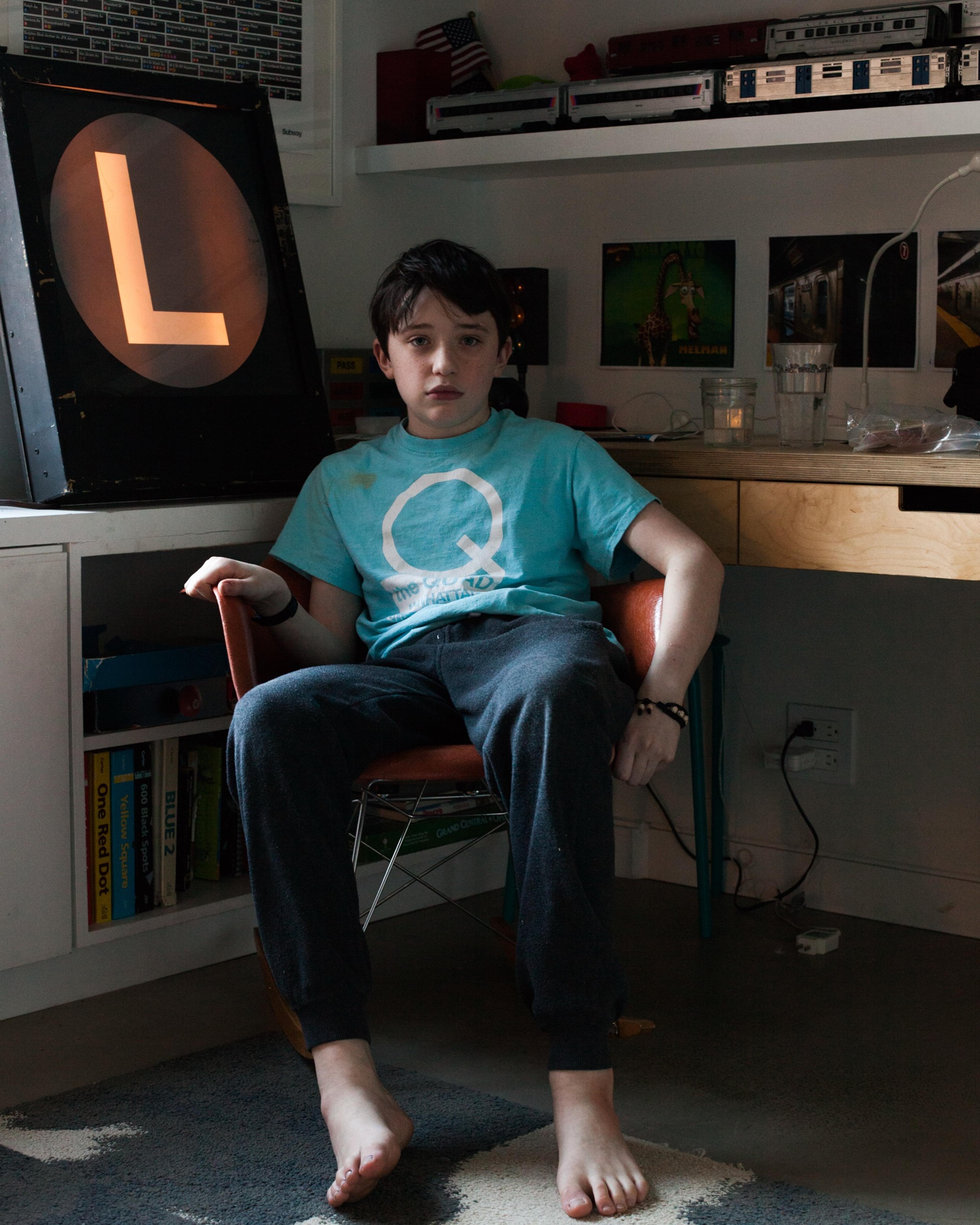 Booker, barefoot in a blue T-shirt, sits in a chair in his room beside a lit-up L train sign, model trains lining the shelves behind him.