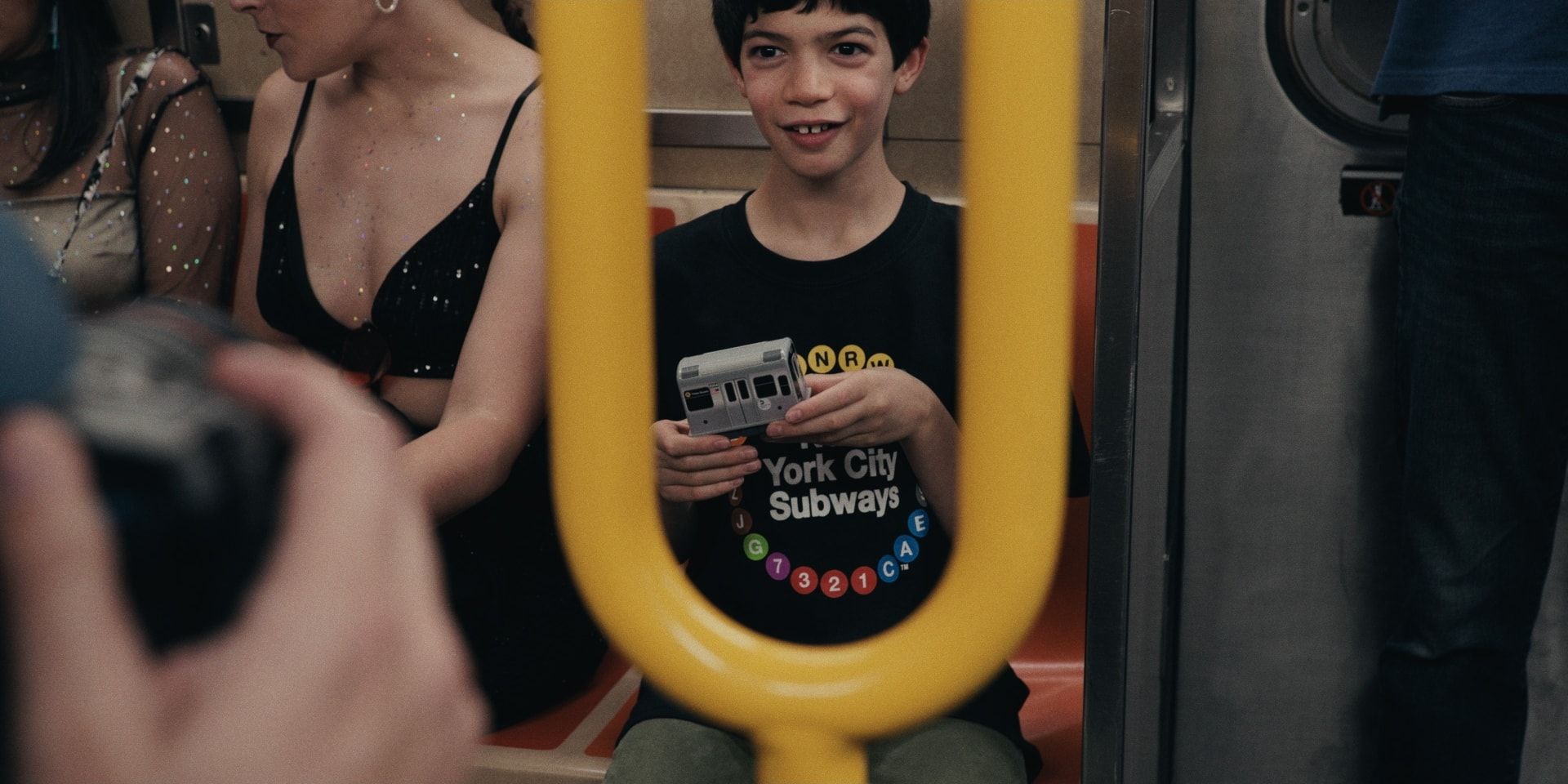 A boy in a New York City Subways T-shirt smiles through a yellow grab handle on a crowded train.