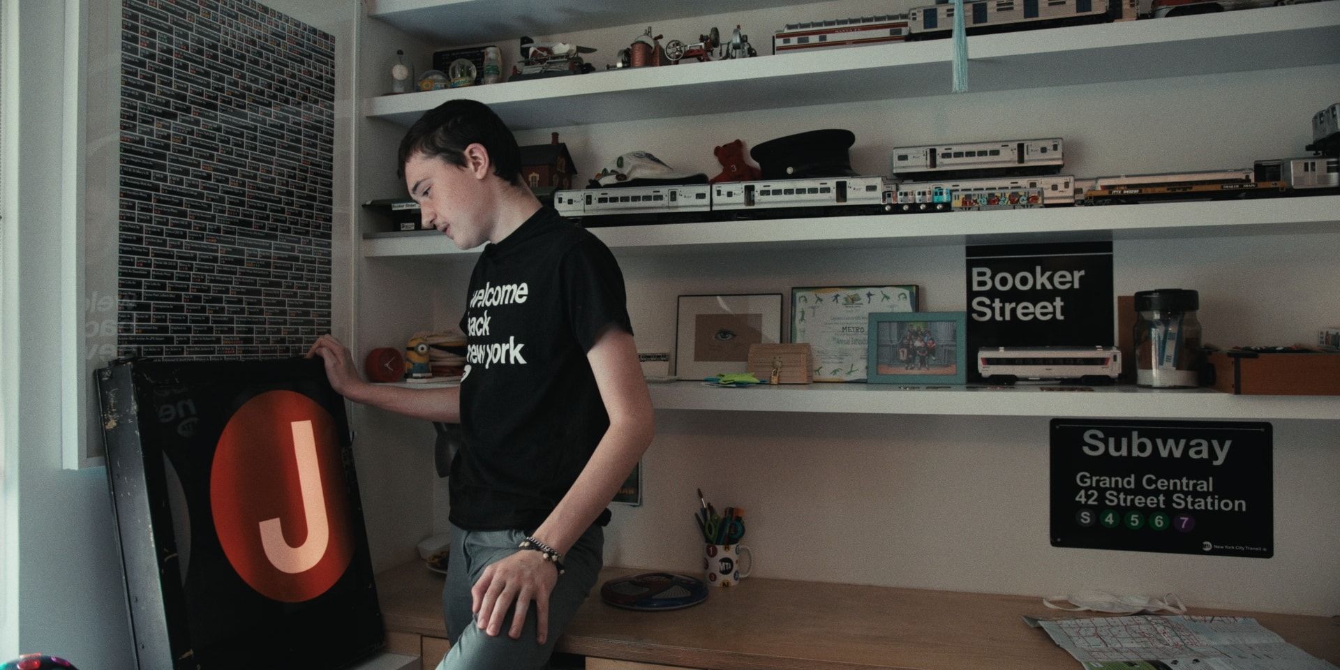 A teenager in a “Welcome Back New York” T-shirt stands beside a J train sign, shelves of model trains and subway signs behind him.