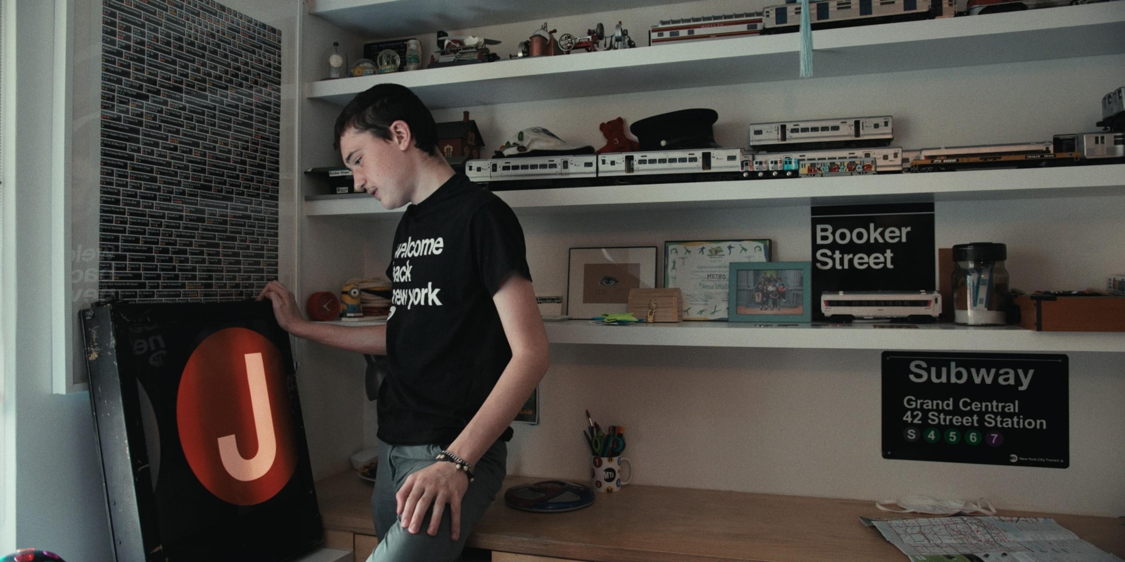 A teenager in a “Welcome Back New York” T-shirt stands beside a J train sign, shelves of model trains and subway signs behind him.