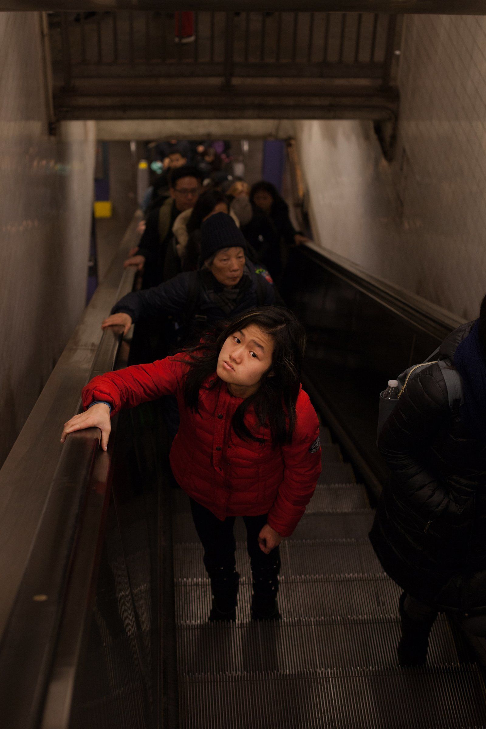 Marley, in a red puffer jacket, looks up as she rides a crowded subway escalator.
