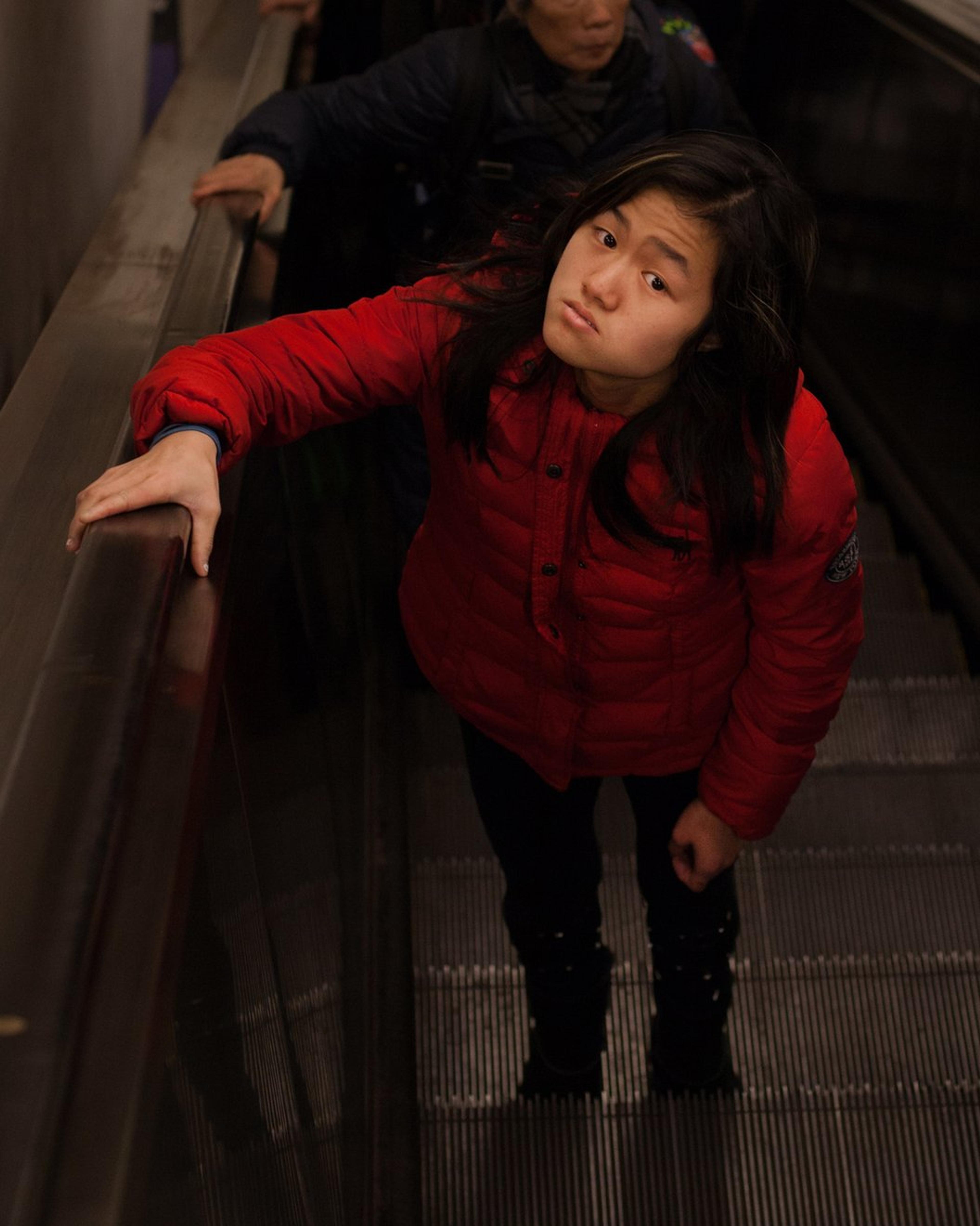 Marley, in a red puffer jacket, looks up as she rides a crowded subway escalator.
