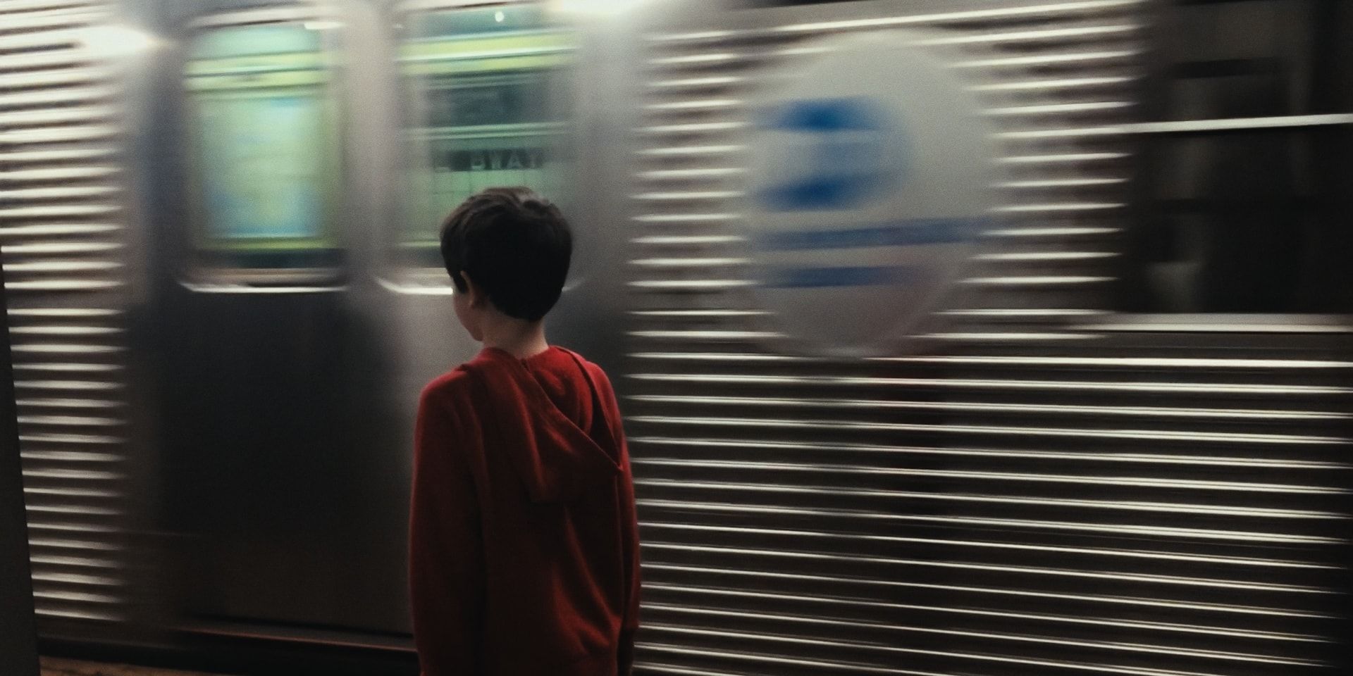 A boy in a red hoodie stands on the platform as a train blurs past.