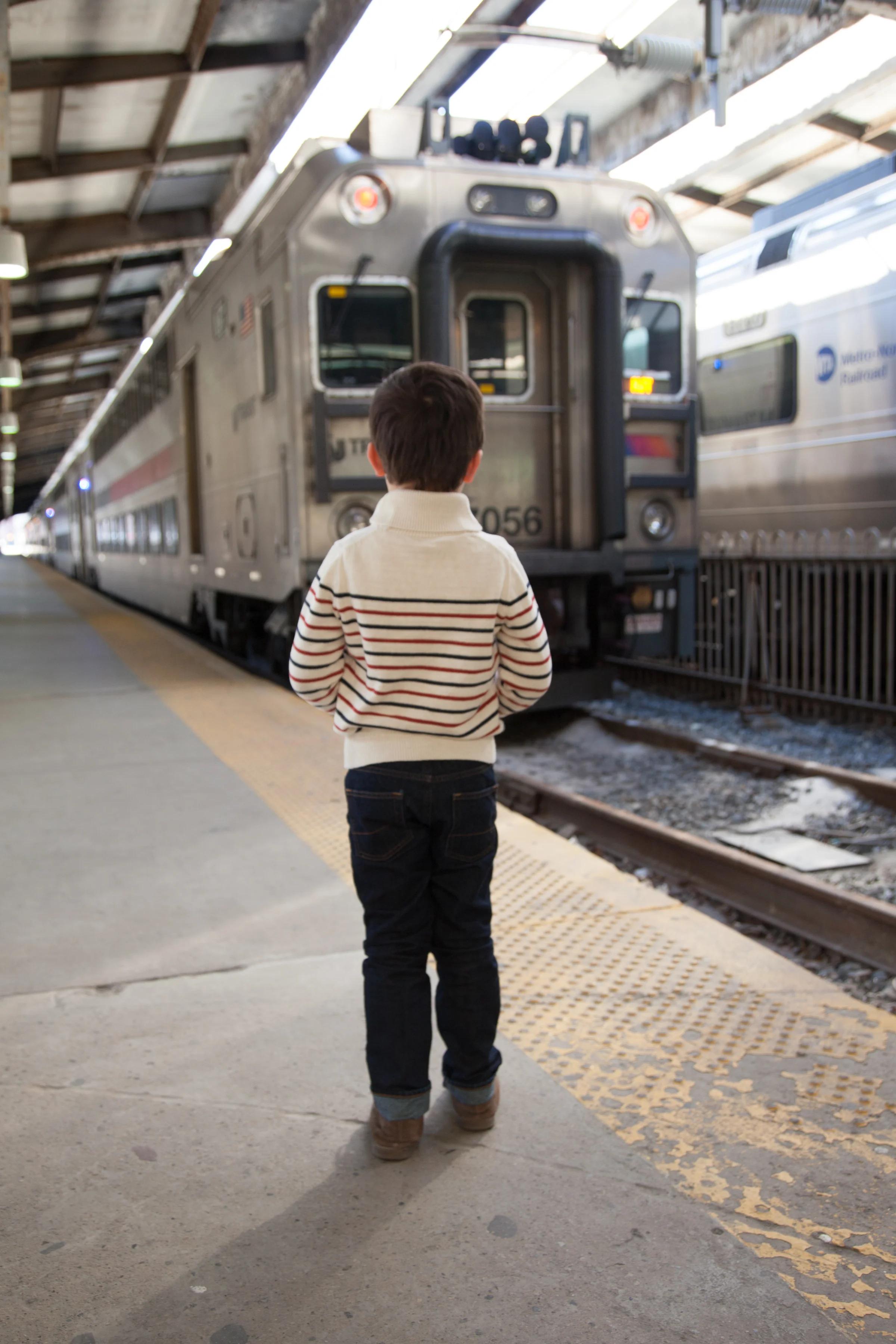 A boy in a striped sweater watches a commuter train from the platform, seen from behind.