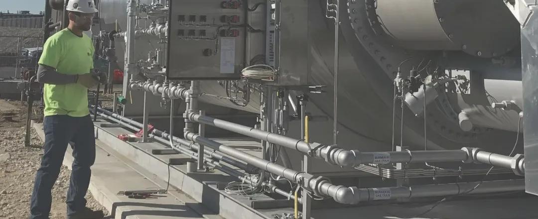 A worker in a hard hat and safety vest beside industrial piping and electrical equipment