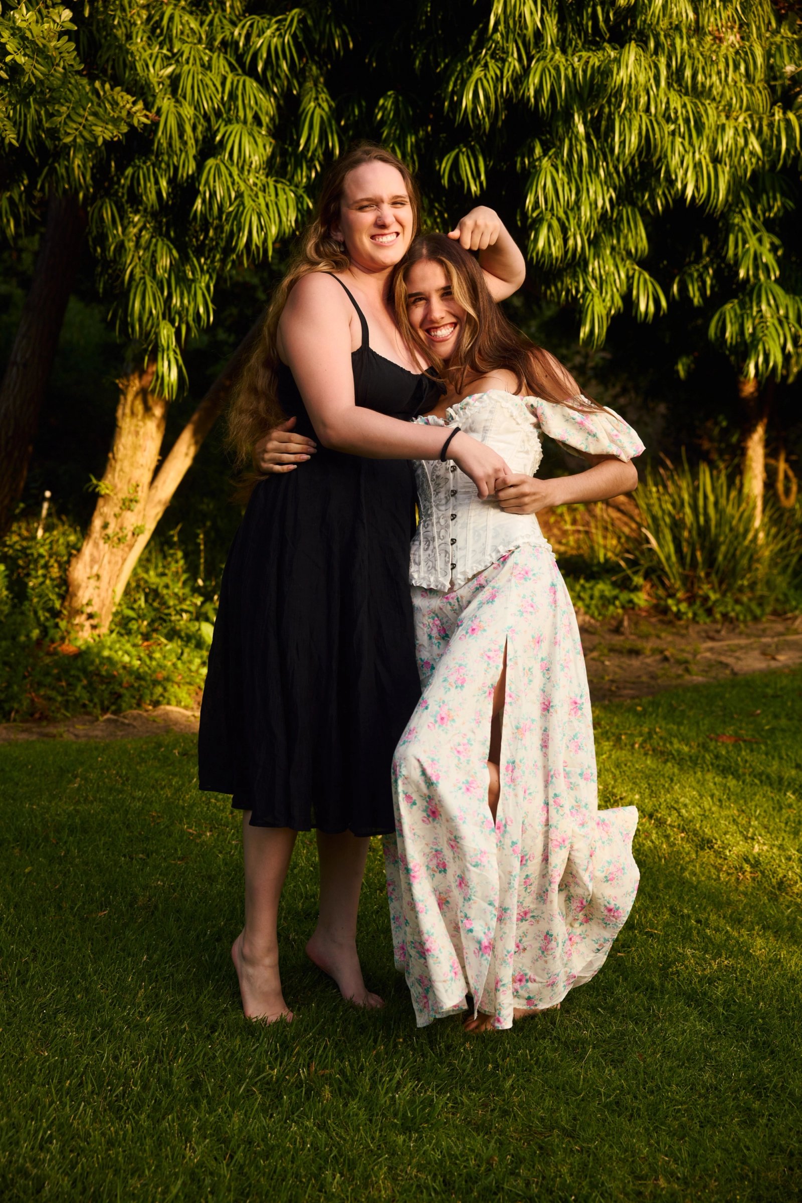 Two people, one in a black dress and one in a white floral dress, embrace on a sunlit lawn beneath leafy trees.