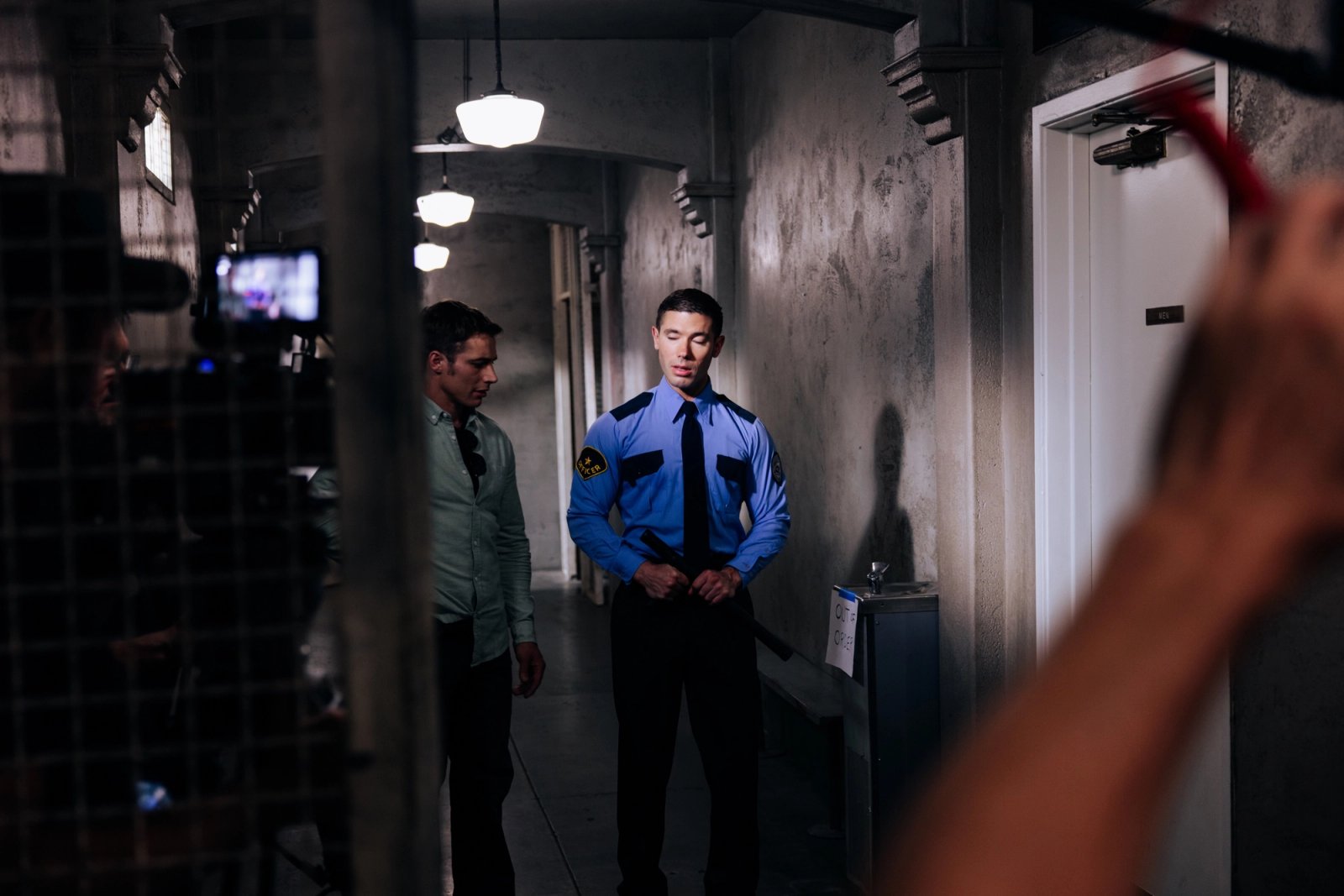 A cinema camera frames two people in a dim concrete hallway, one wearing a blue uniform under an overhead light.