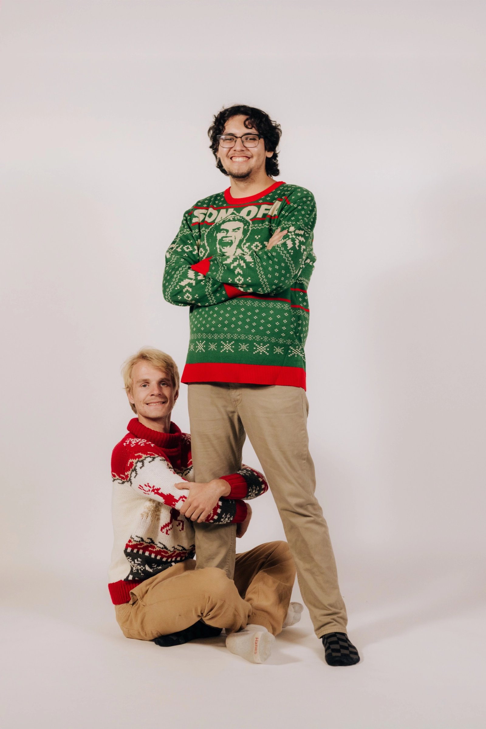 Two people in green and red holiday sweaters pose on a white studio backdrop, one standing and one kneeling.