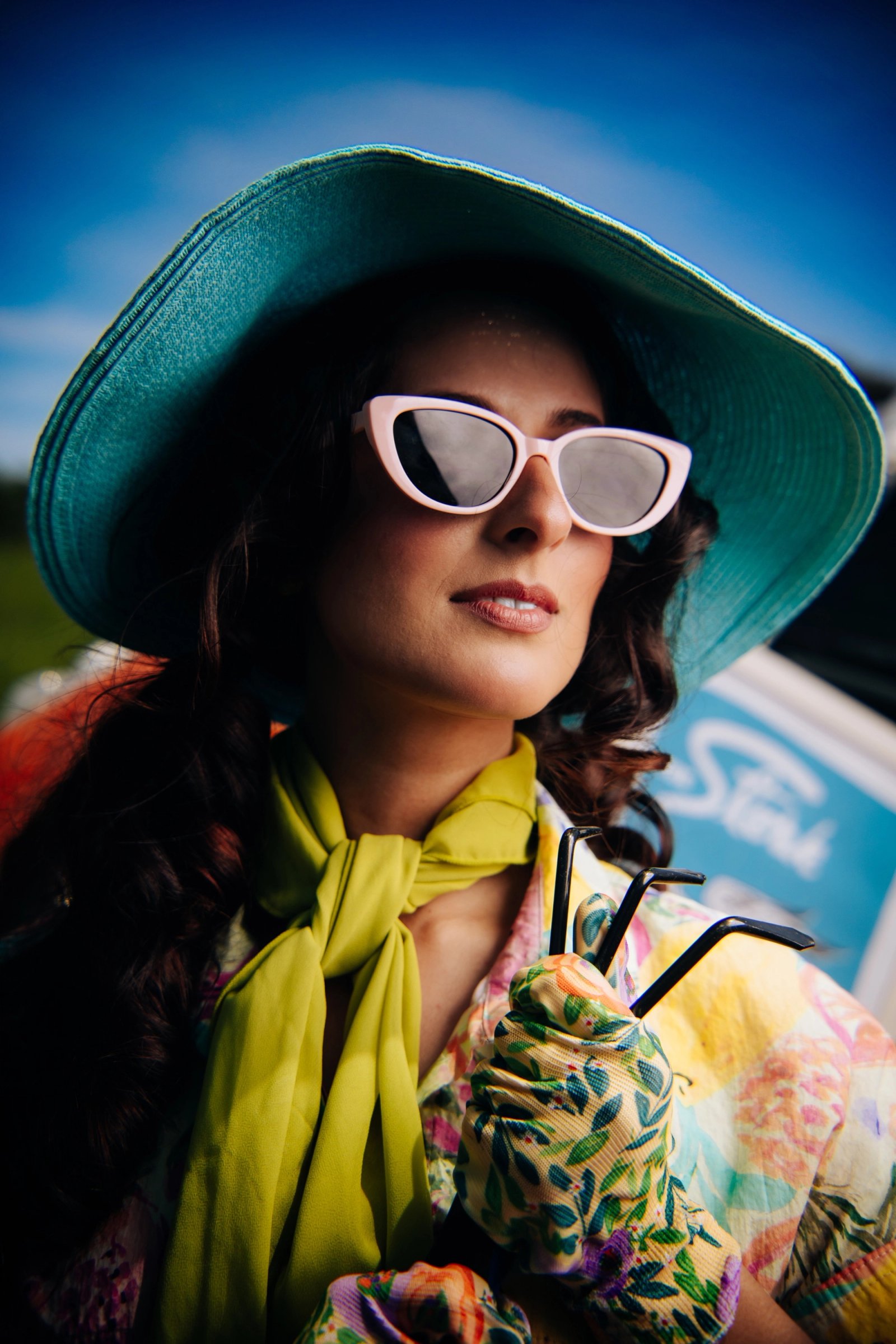 A close portrait of a person in a teal wide-brimmed hat, white sunglasses, and a lime scarf against a vivid blue sky.