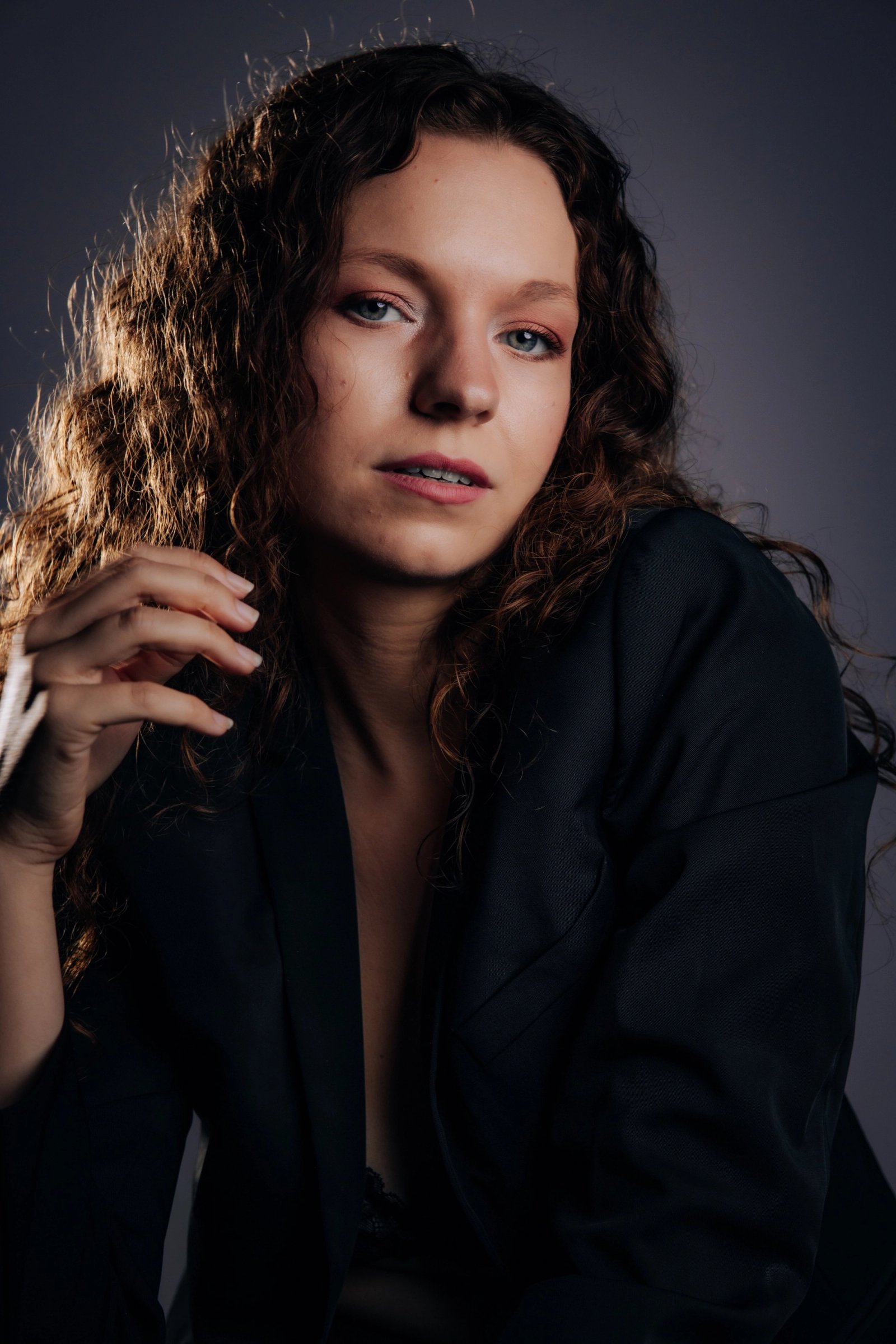 A close studio portrait of a curly-haired person in a black blazer with one hand raised near their face against a gray backdrop.