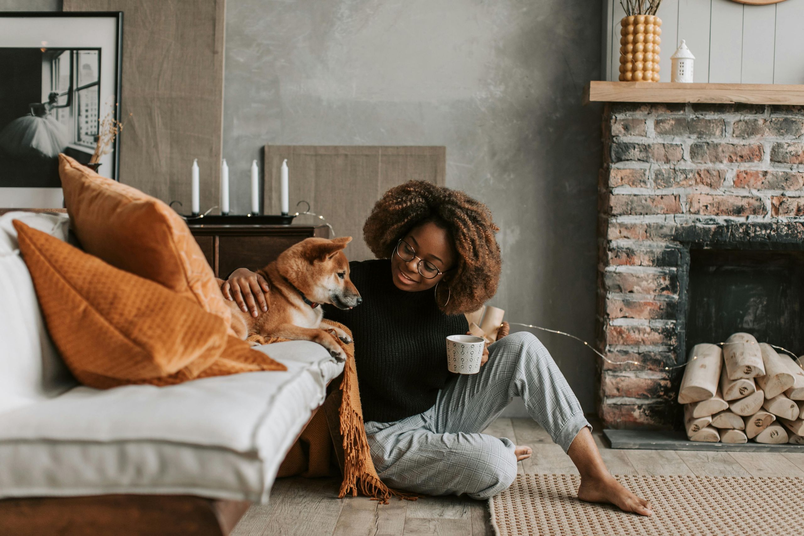 A woman sits with a cup of tea in front of the couch, petting her dog.
