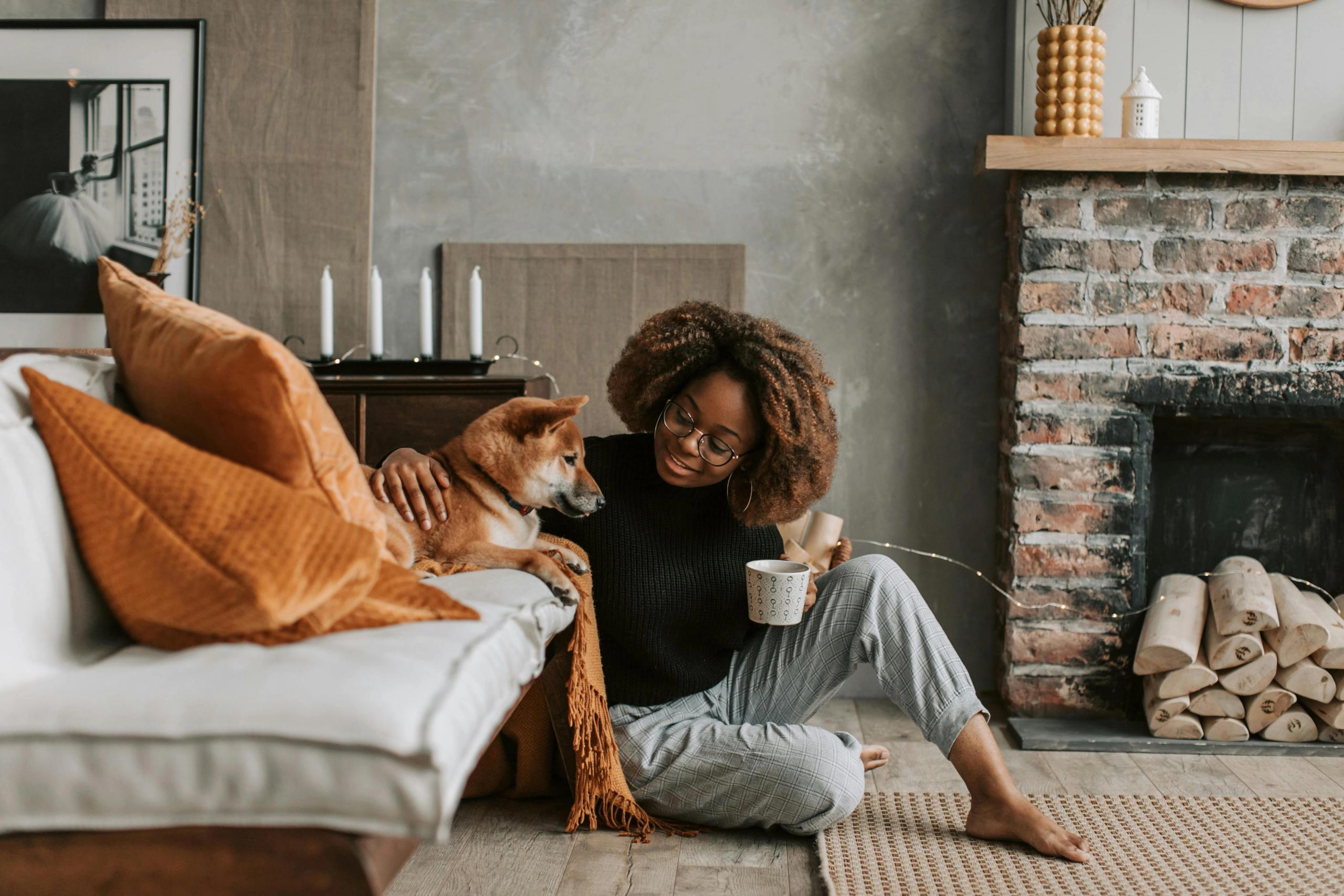 A woman sits with a cup of tea in front of the couch, petting her dog.