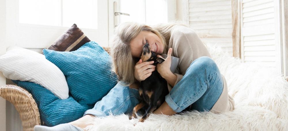 A smiling woman hugs a small black and tan dog on a fluffy white couch.