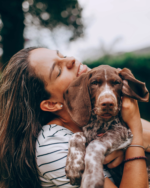 A smiling woman with closed eyes hugs a brown and white spotted puppy looking at the camera.