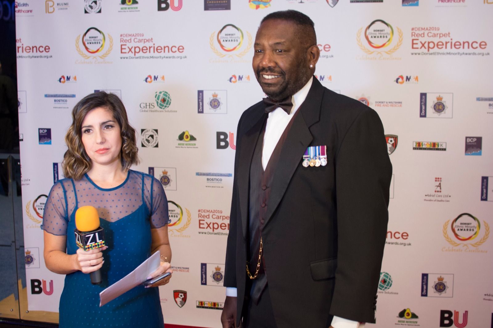 A presenter with a microphone and a guest in a dinner suit on the red carpet