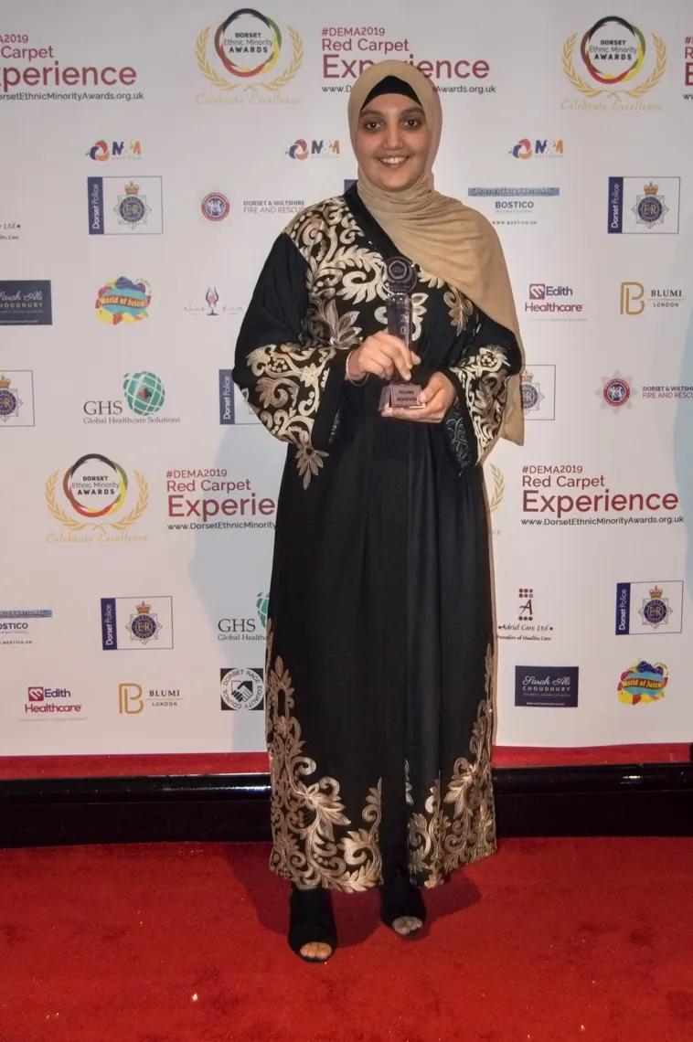 A winner in an embroidered abaya holding her trophy on the red carpet