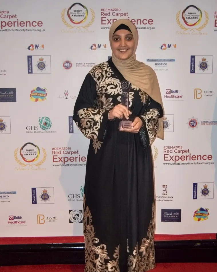 A winner in an embroidered abaya holding her trophy on the red carpet